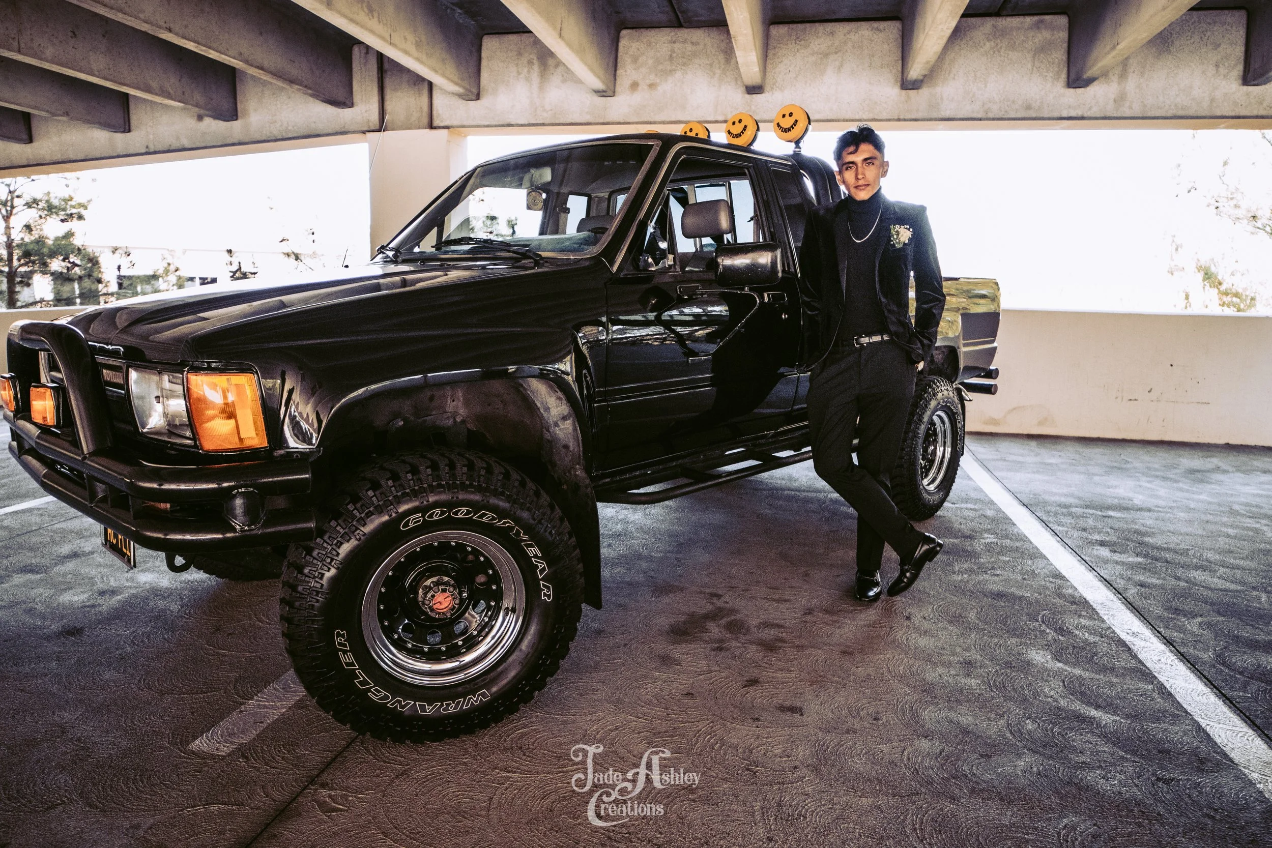 A young man dressed in black standing in an underground parking garage next to a black pickup truck decorated with smiley face lights on the roof.