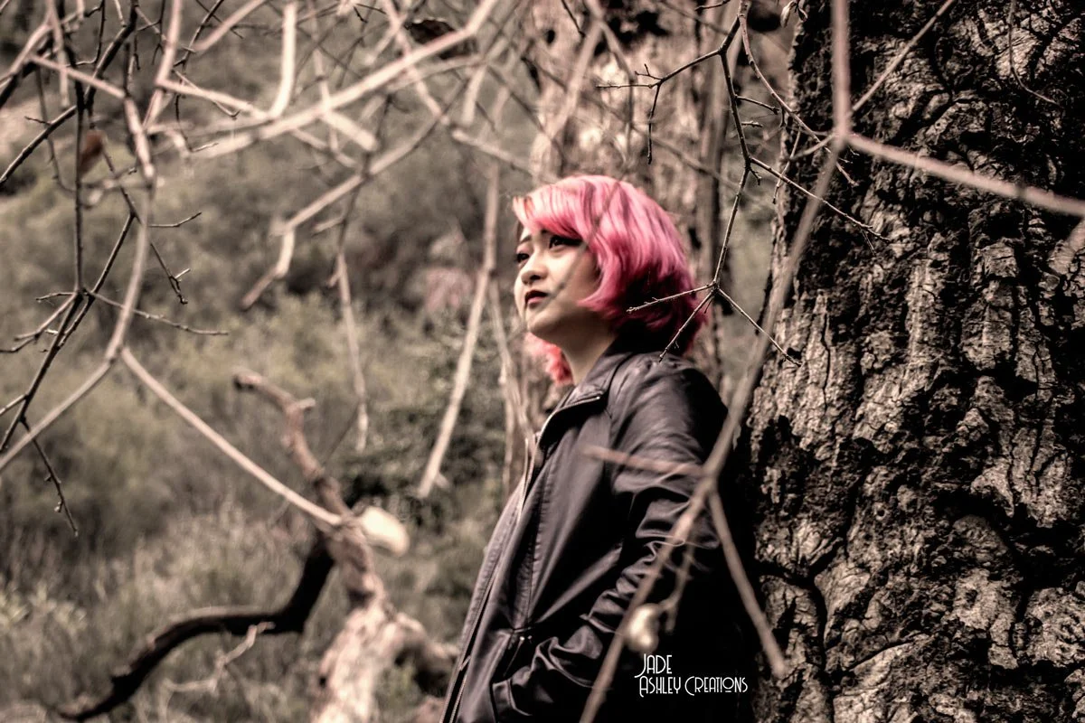A woman with pink hair leaning against a large tree trunk in a wooded area, looking to the side.