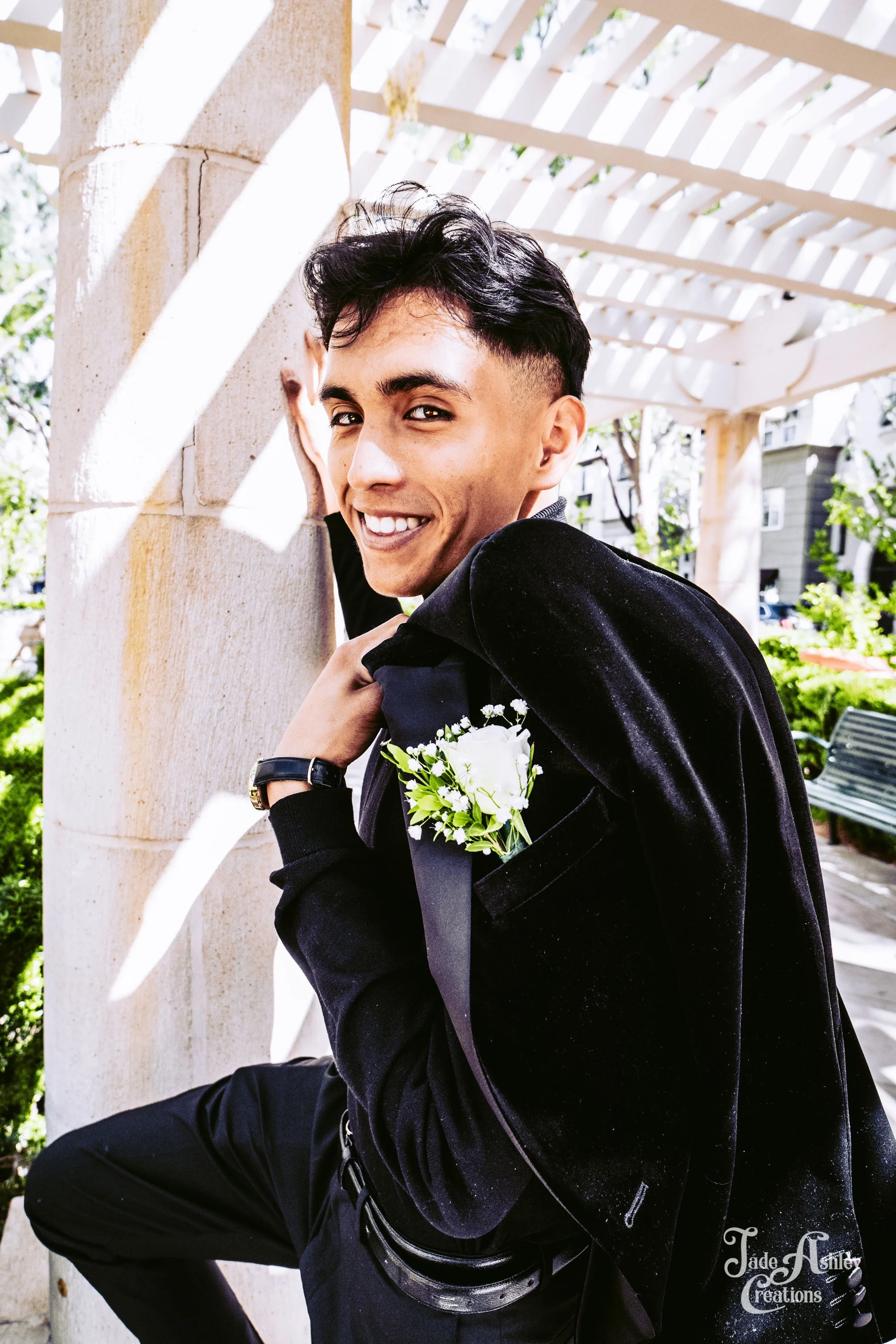 Young man in a black blazer with a white boutonniere, smiling and posing outdoors near a wooden structure.
