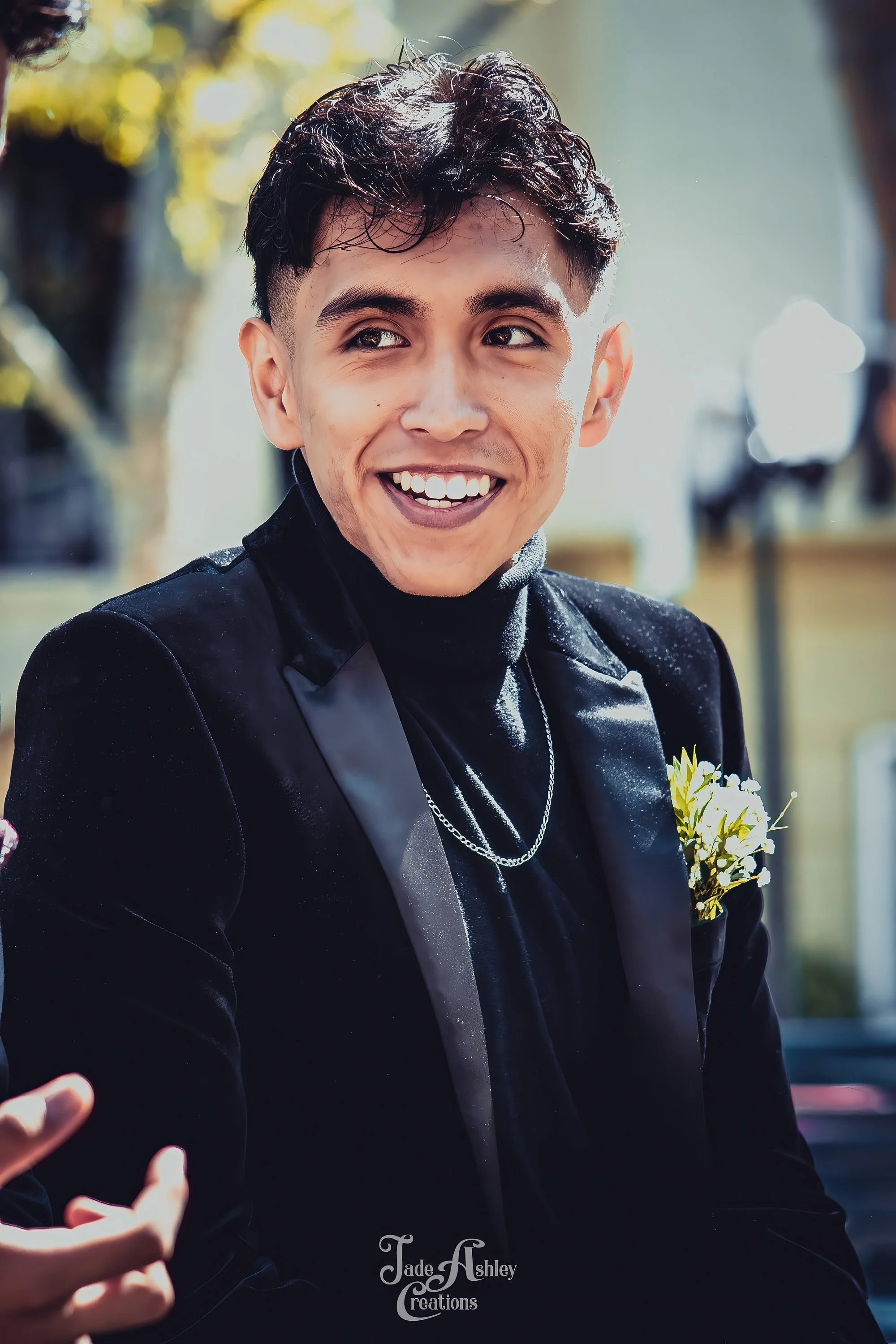 A young man with dark, wavy hair, smiling and looking to the side, dressed in black formal attire with a flower boutonniere on his jacket, outdoors with trees and sunlight in the background.