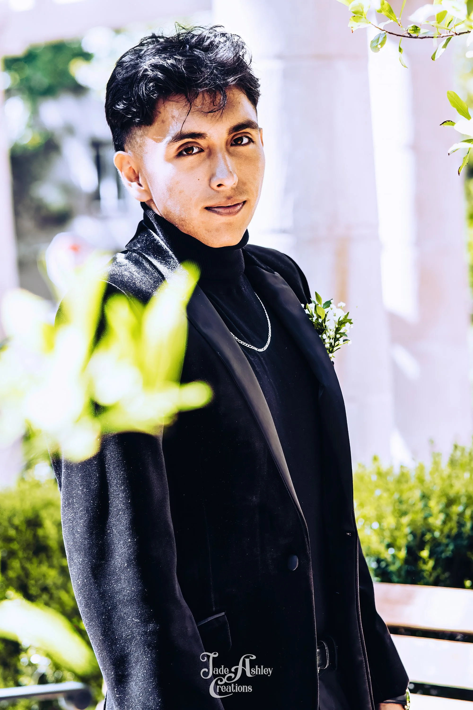 A young man with dark hair is wearing a black velvet tuxedo jacket over a black turtleneck. He is outdoors, with greenery and a white building in the background, and is looking at the camera with a slight smile.