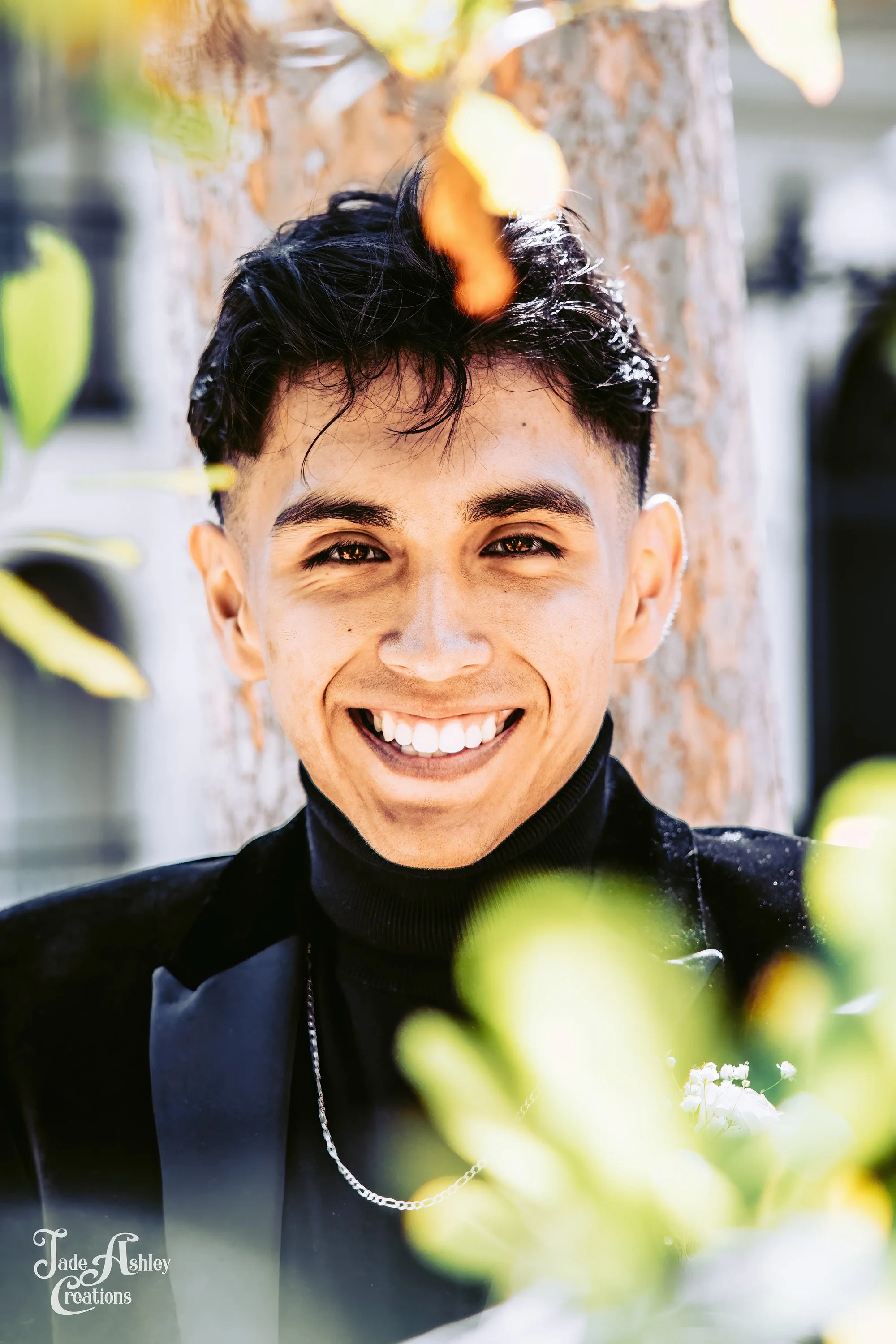 A young man smiling brightly outdoors, wearing a black turtleneck and black blazer, with a chain necklace, standing near a tree with green leaves.