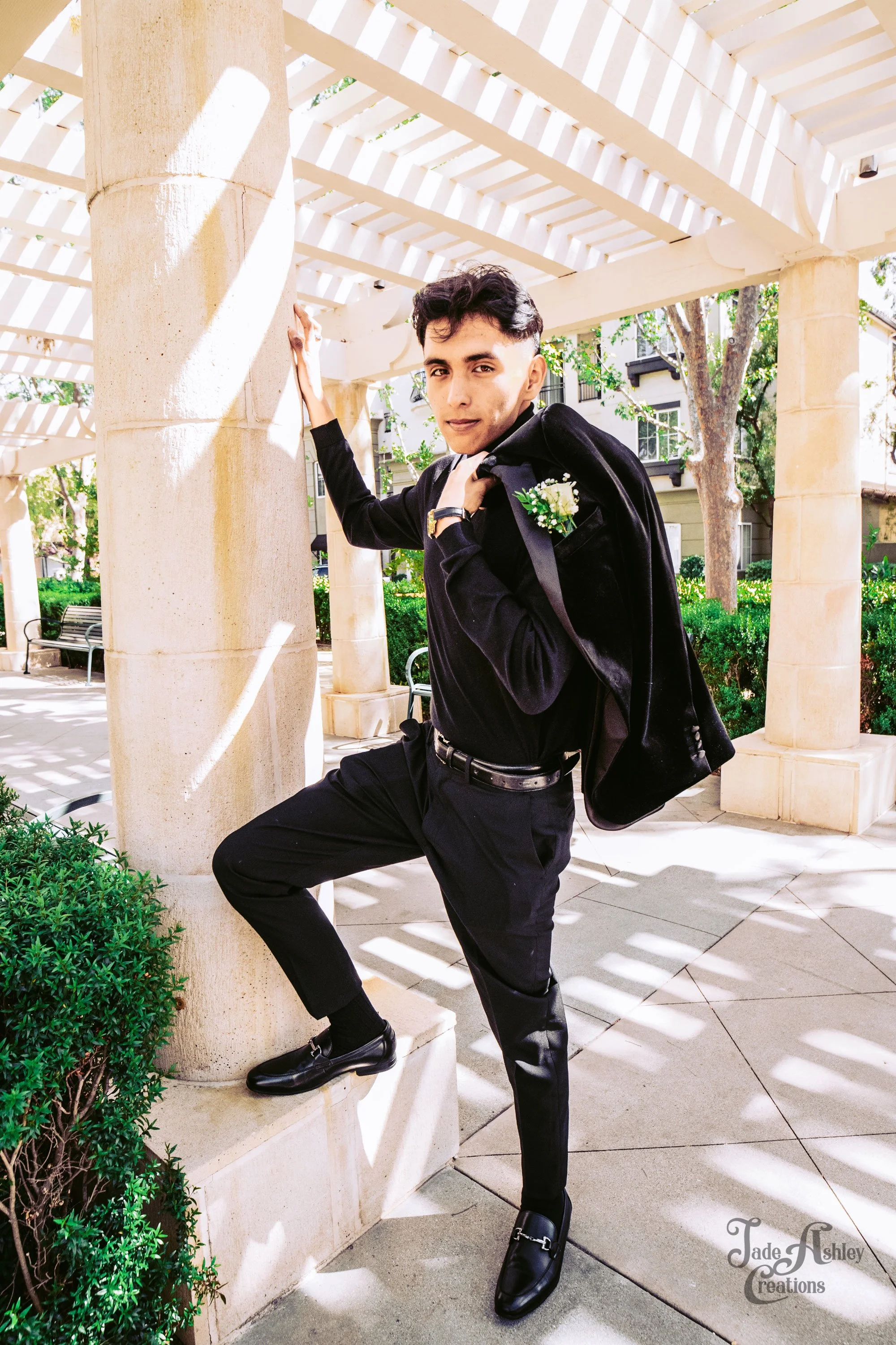 A young man in a black suit standing outdoors under a white pergola, with one foot on a stone pillar, holding a black jacket over his shoulder, and wearing a boutonniere.