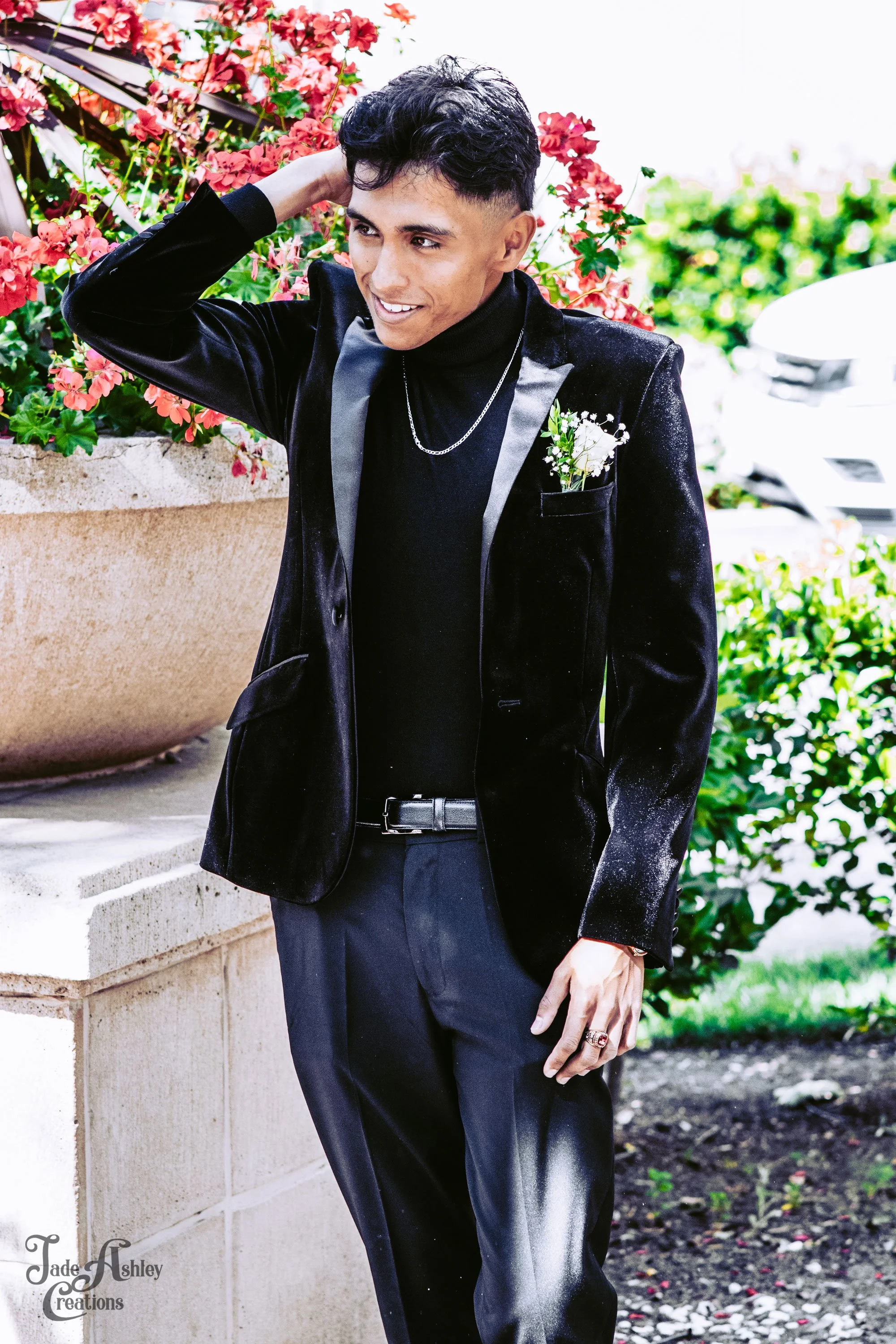A young man with black hair dressed in a black tuxedo jacket, black turtleneck, and dress pants, standing outdoors near a flower planter with pink flowers. He is smiling and has his left hand resting on his head.