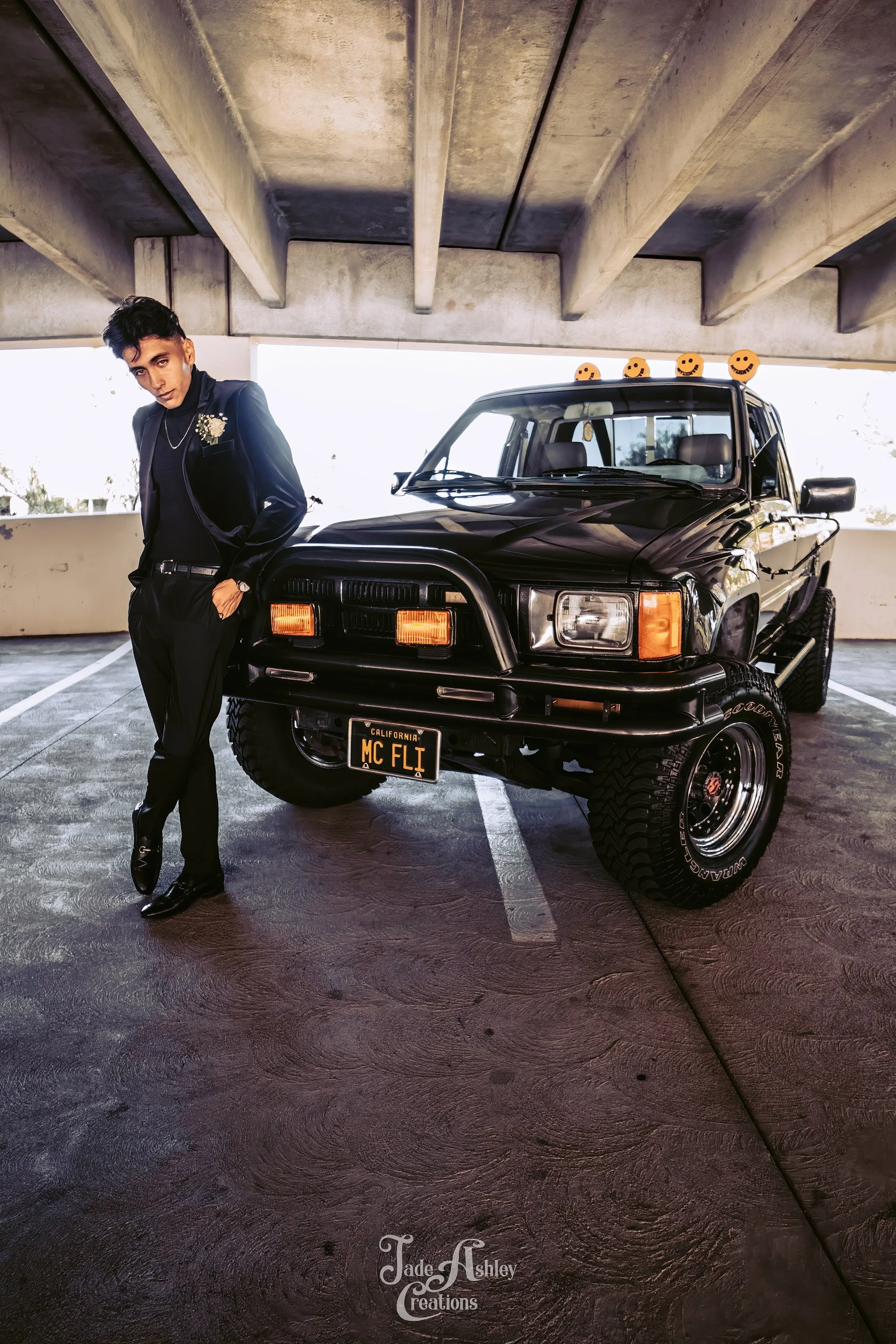 A young man in a black suit with floral boutonniere standing next to a black off-road vehicle with a California license plate. The vehicle has pumpkin-shaped decorations on the roof.