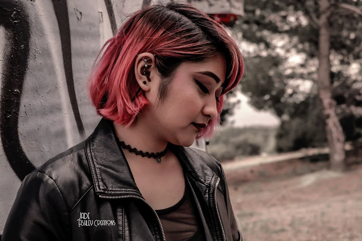 A woman with short pink hair and multiple ear piercings, wearing a black leather jacket and choker necklace, stands outdoors near a graffiti wall and trees.