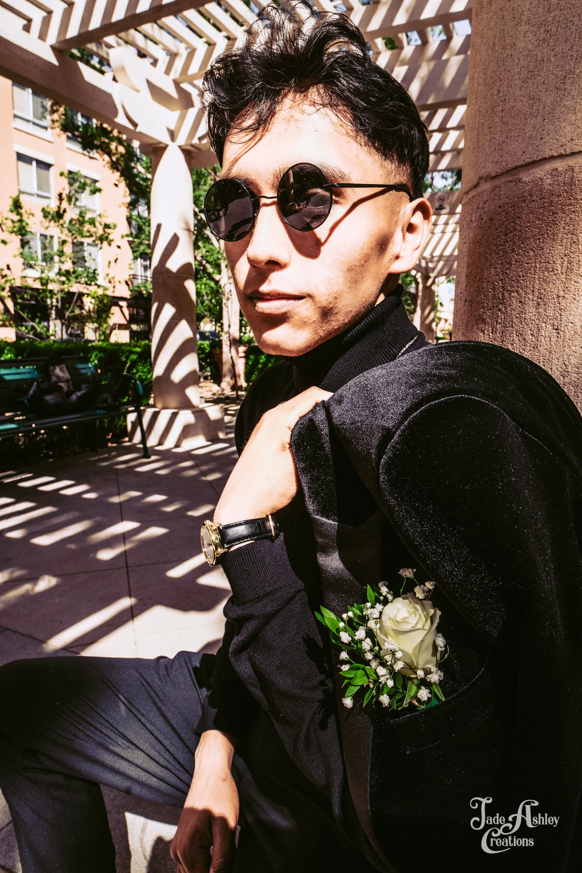 A person sitting outdoors, wearing sunglasses, a black outfit with a boutonniere, and a gold watch, with a decorative structure casting shadows behind them.