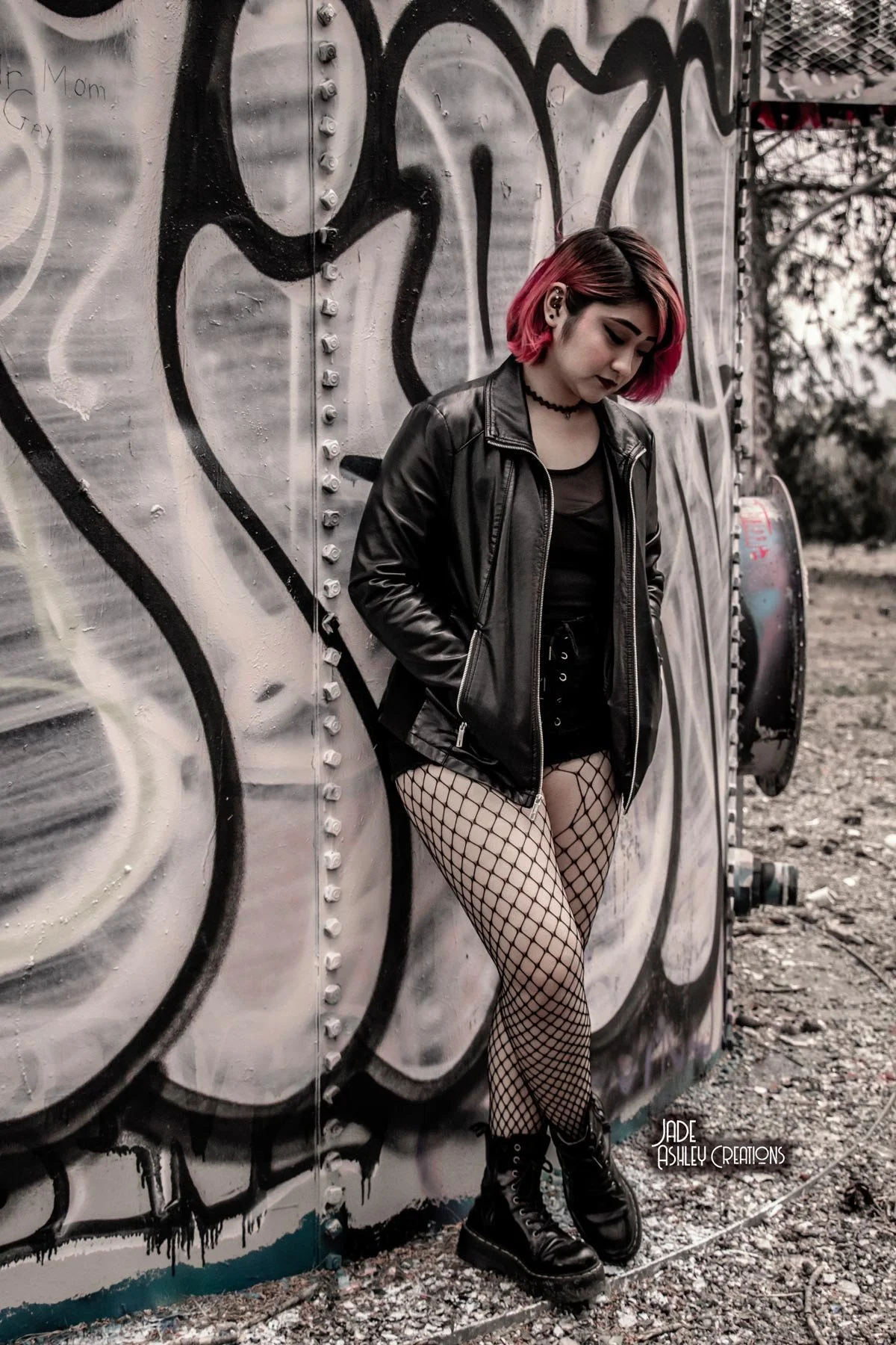 Young woman with pink hair, wearing a black leather jacket, fishnet stockings, and black combat boots, leaning against a graffiti-covered wall outdoors.