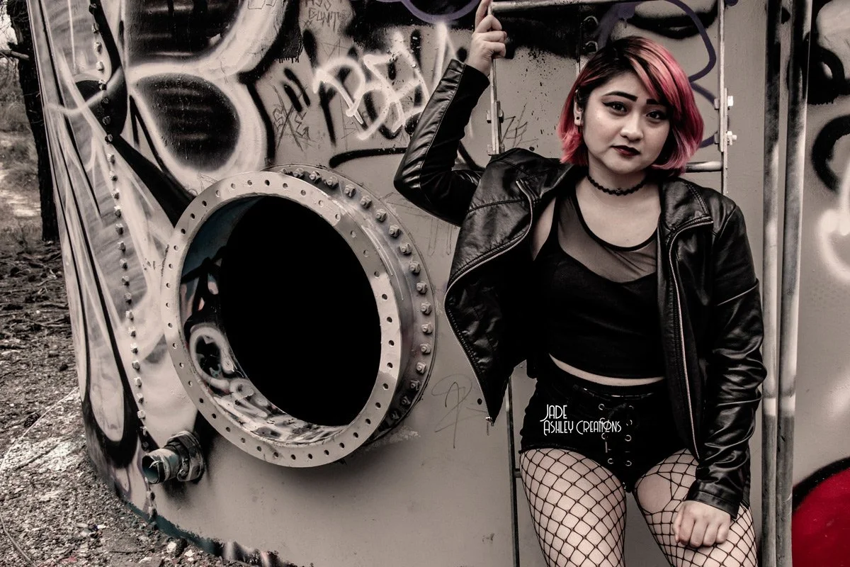 A young woman with pink and blonde hair wearing a black leather jacket, black crop top, fishnet stockings, and dark lipstick, standing in front of a graffiti-covered laundromat dryer with the door open.