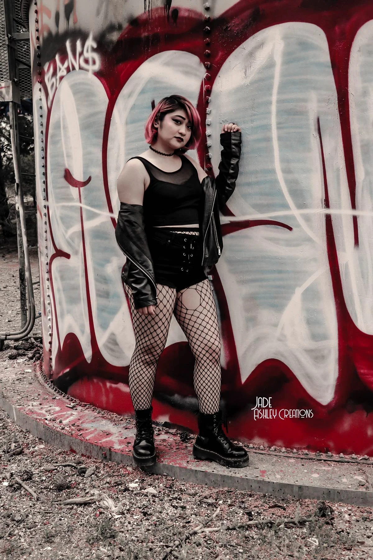 A young woman with pink hair and dark makeup standing next to a graffiti-covered metal structure outdoors, wearing a black crop top, fishnet tights, black boots, and a black leather jacket.