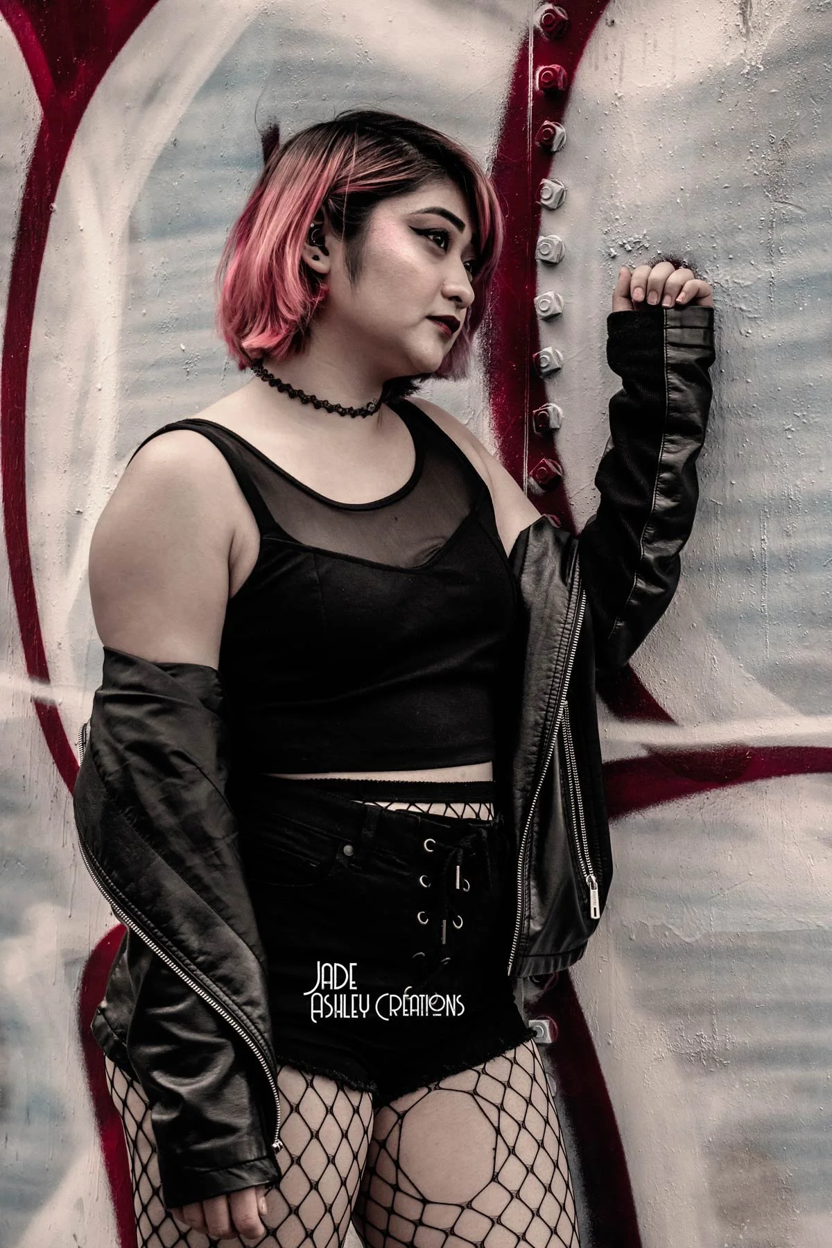 A young woman with pink and black hair, wearing a black tank top, fishnet stockings, and a black leather jacket, standing against a graffiti-covered wall.