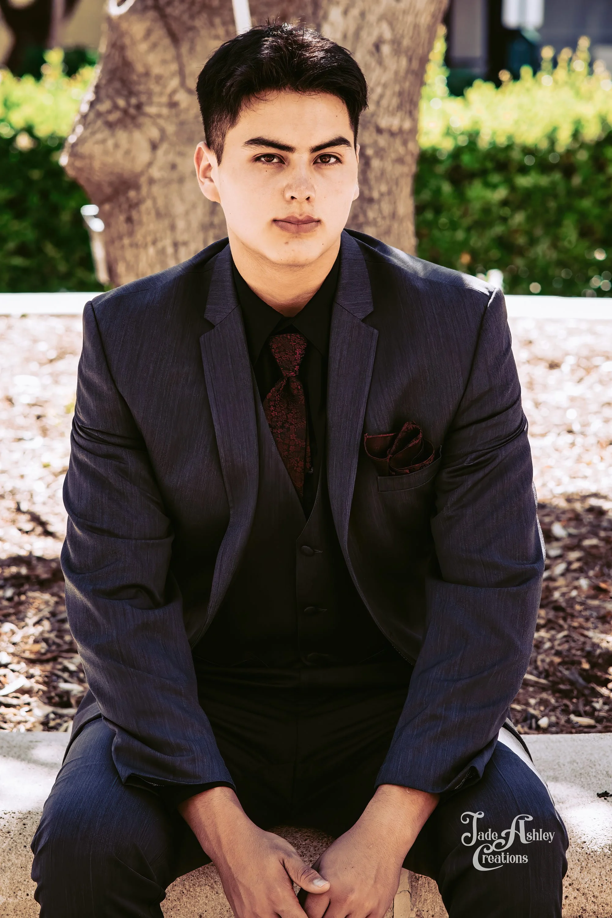 Young man wearing a dark suit with a red tie and pocket square, sitting outdoors in front of a tree, looking at the camera.