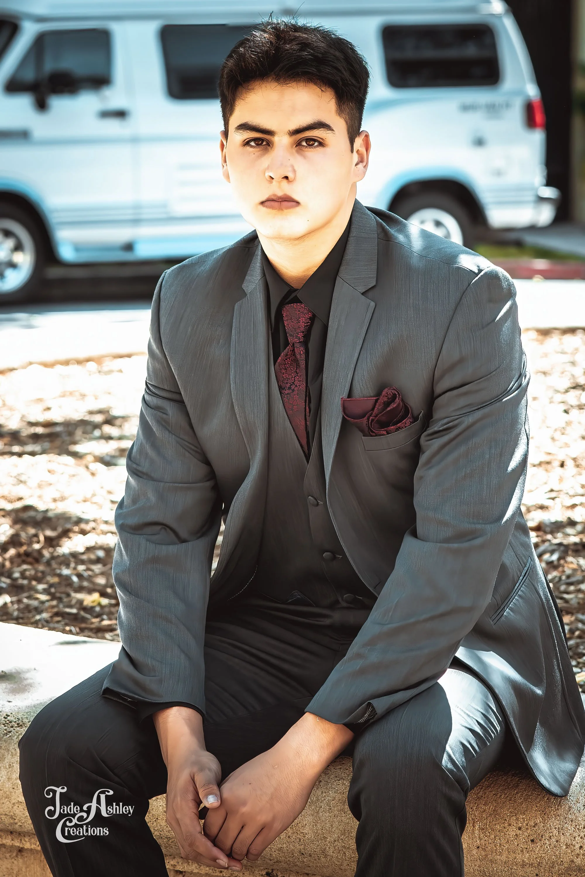 A young man in a gray suit sitting on a stone ledge outdoors with a light blue van in the background.