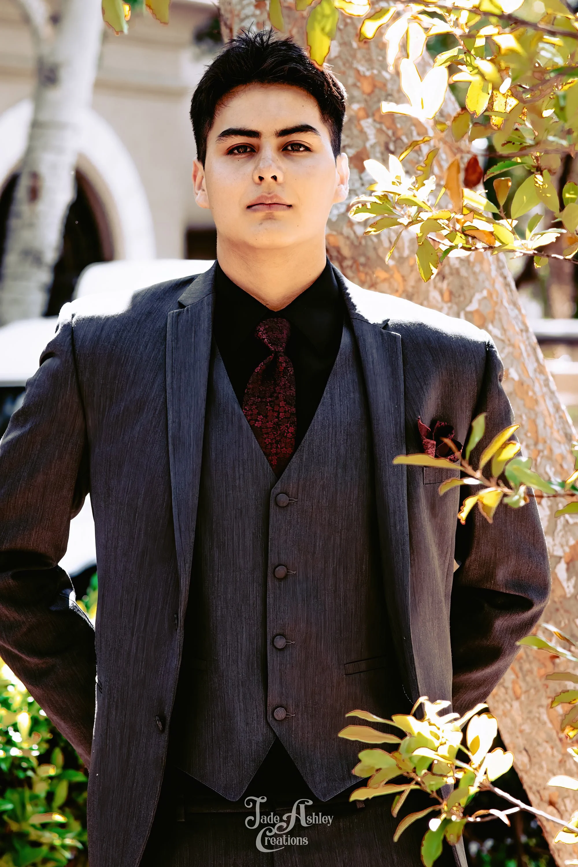 A young man dressed in a dark gray suit with a matching vest, black shirt, and dark patterned tie, standing outdoors near a tree with sunlight filtering through the leaves.