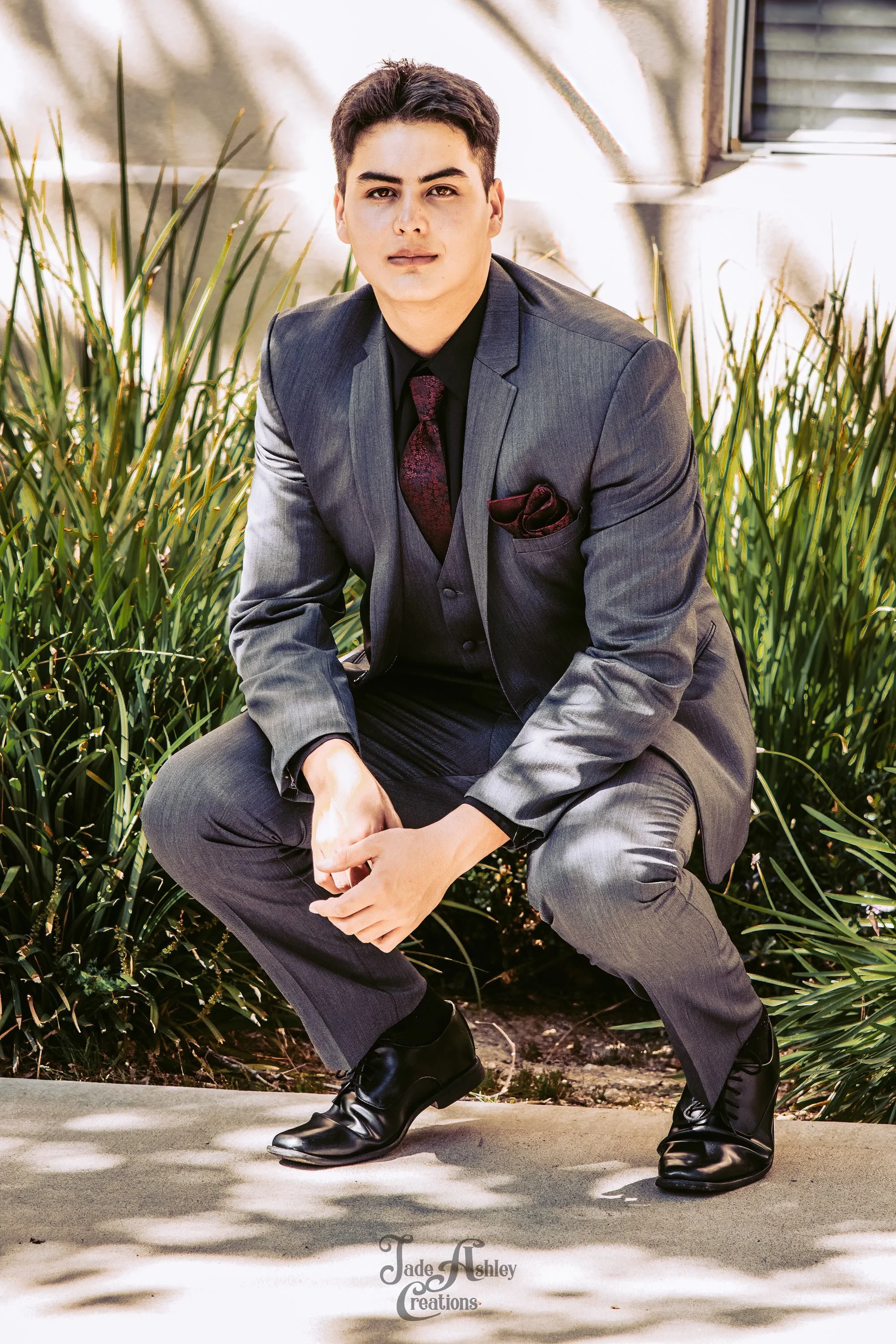 A young man in a gray suit with a black shirt and maroon tie, squatting outdoors on a sidewalk with greenery and sunlight in the background.