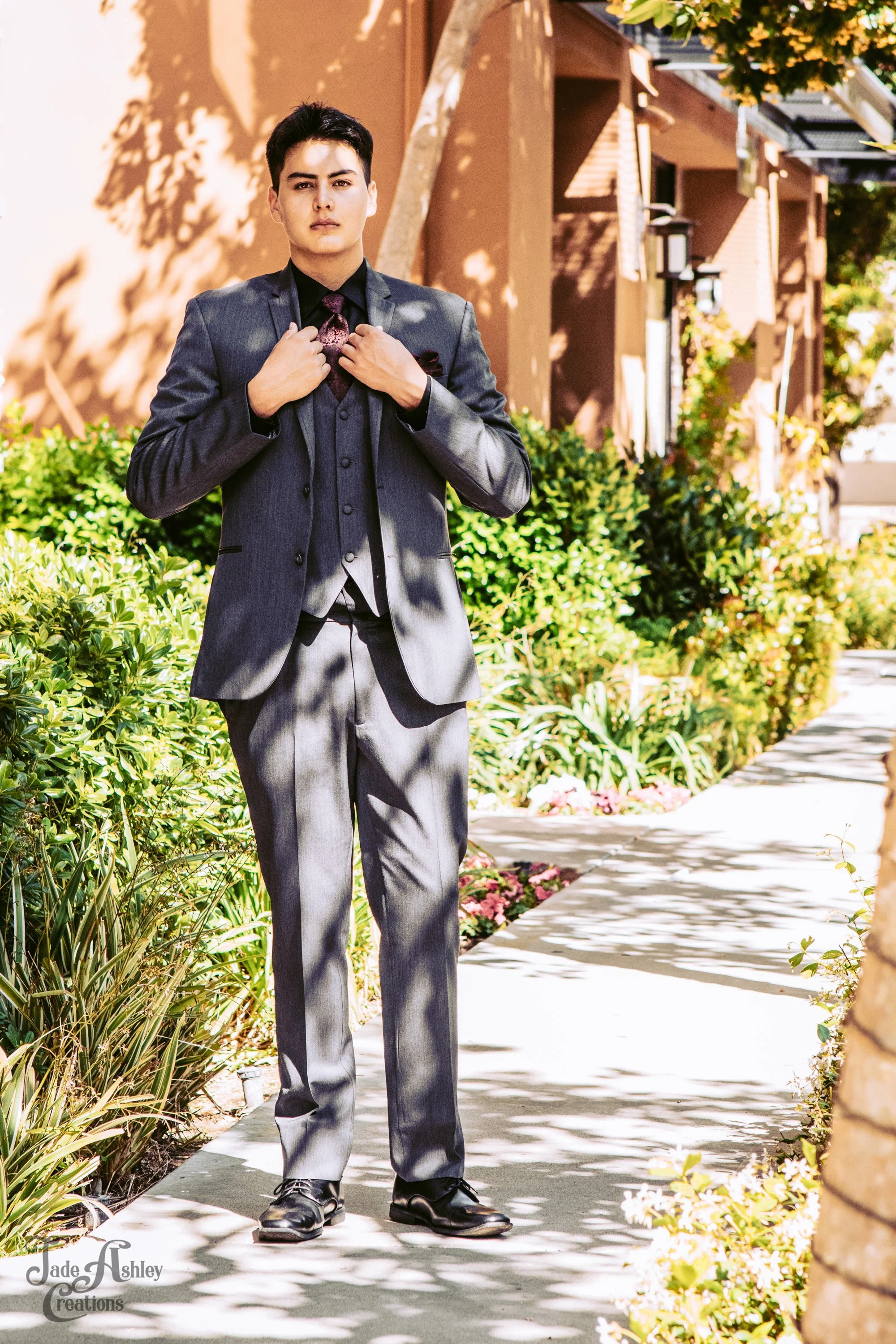 A young man in a gray suit standing on a sidewalk in front of a building with lush greenery and flowers, adjusting his tie.