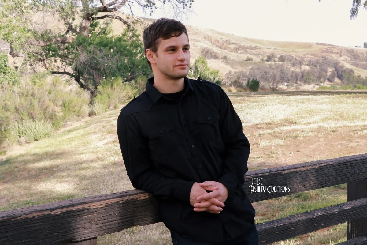 A young man in black clothing leans against a wooden fence outdoors, with rolling hills, trees, and open land in the background.