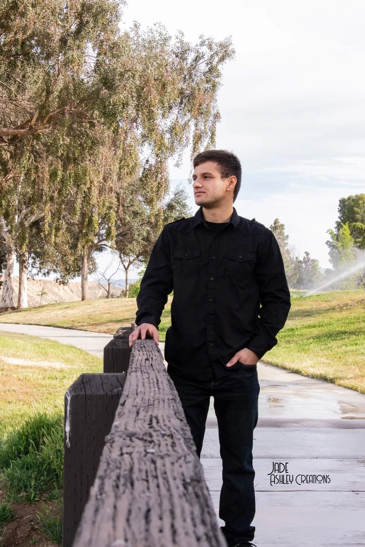 A young man in a black shirt and pants leaning on a wooden park bench in a green outdoor park with trees and a cloudy sky.