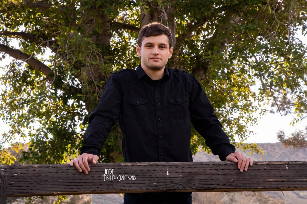 A young man with brown hair and fair skin wearing a black button-up shirt, standing outdoors behind a wooden railing with greenery in the background.