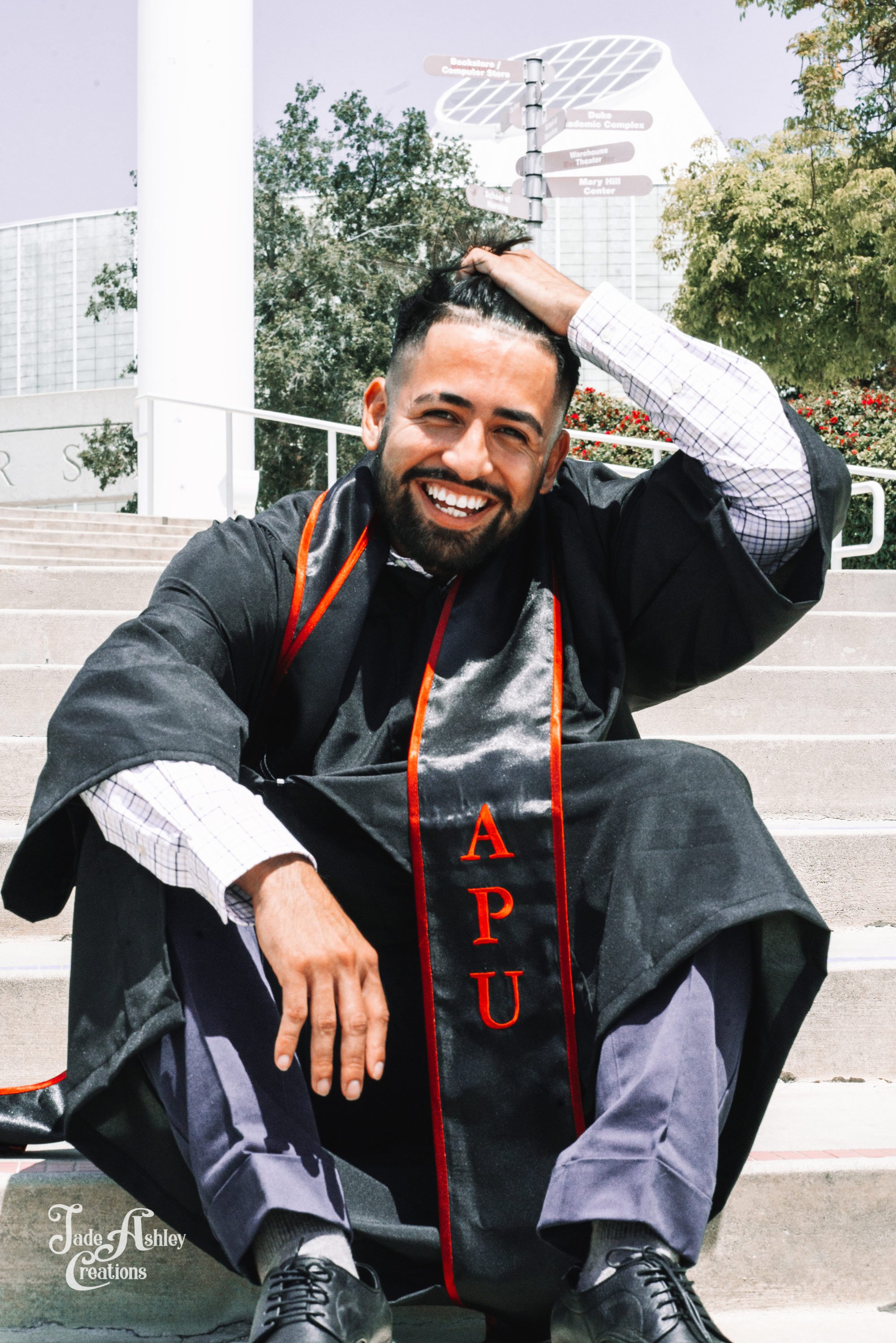 A man wearing a graduation gown and stole with red letters spelling 'APU' sitting on steps outdoors, smiling and touching his hair.