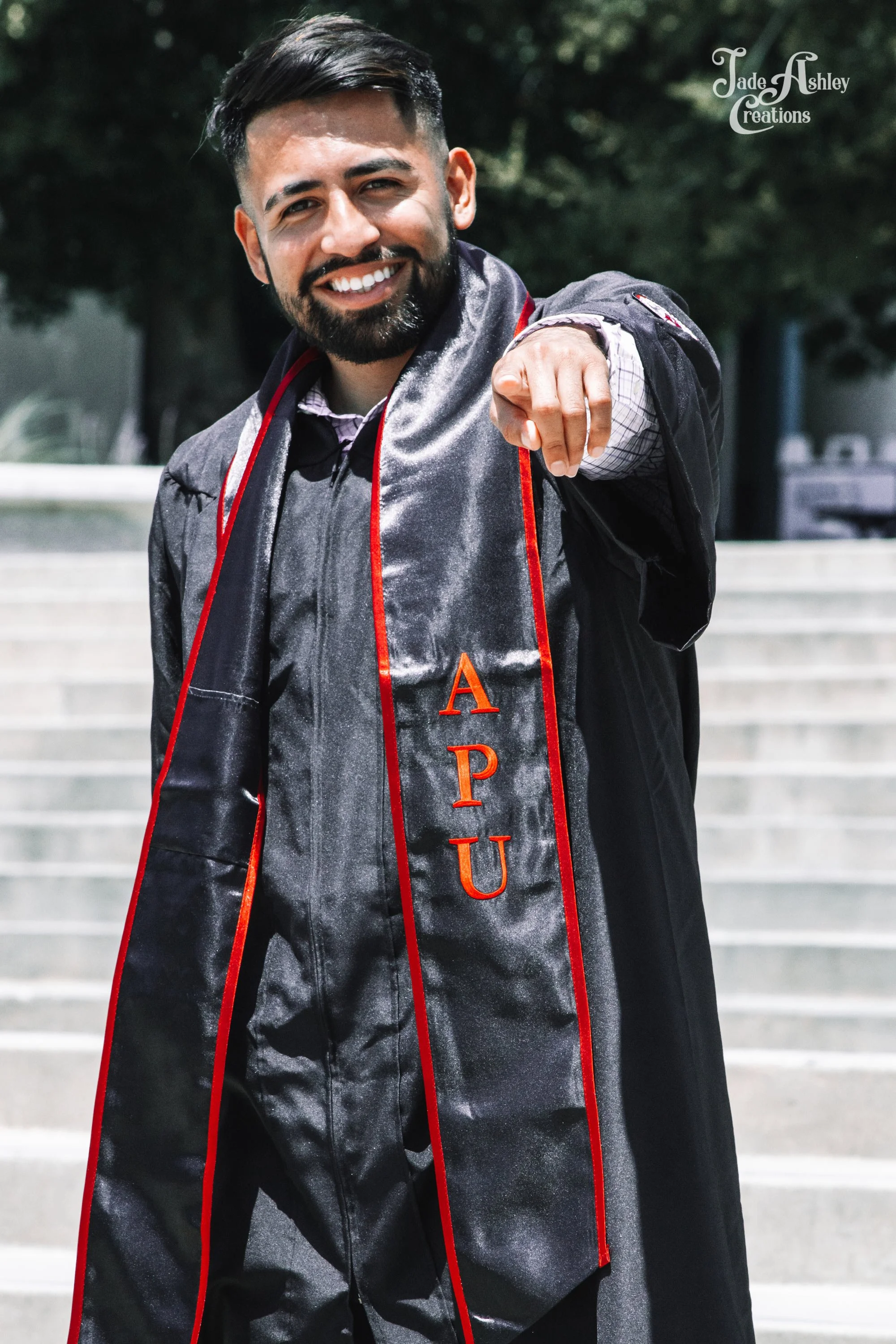 A man in a graduation gown pointing at the camera, wearing a sash with the letters A P U, smiling, outdoors with stairs and trees in the background.