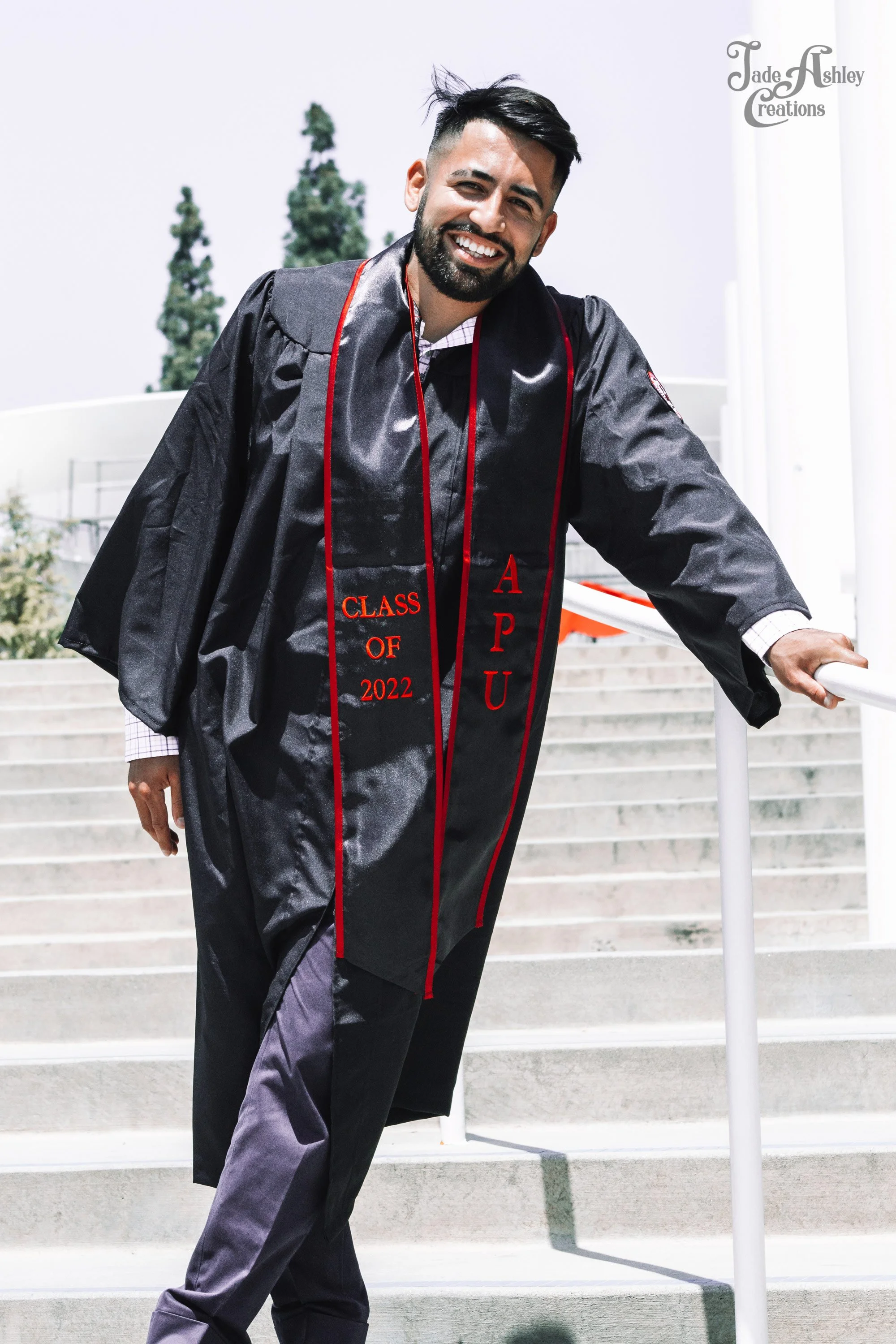 A man in graduation gown and stole on stairs outdoors, smiling at camera, with trees in background.