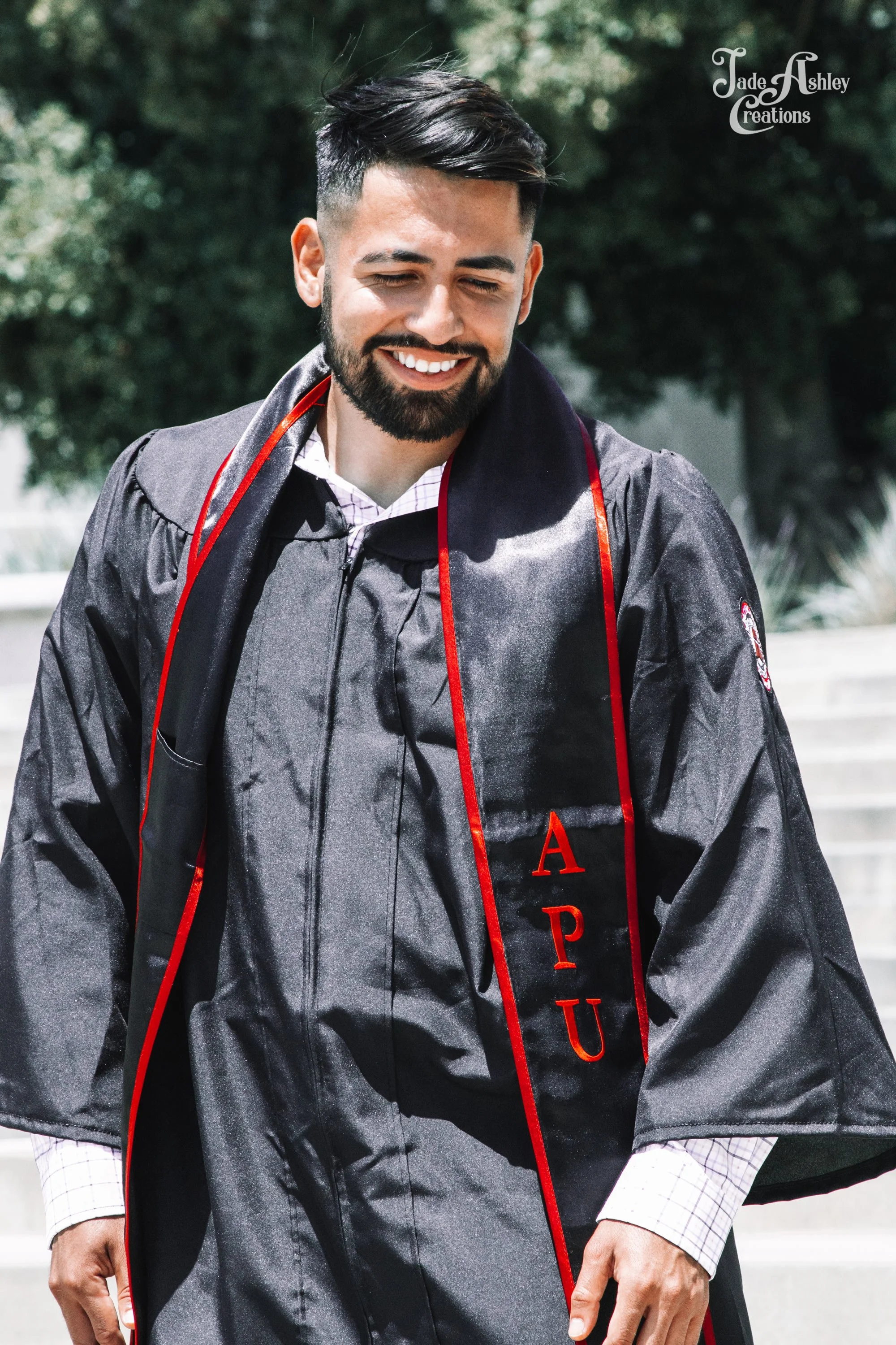 A man in a graduation gown smiling outdoors, with the Greek letters 