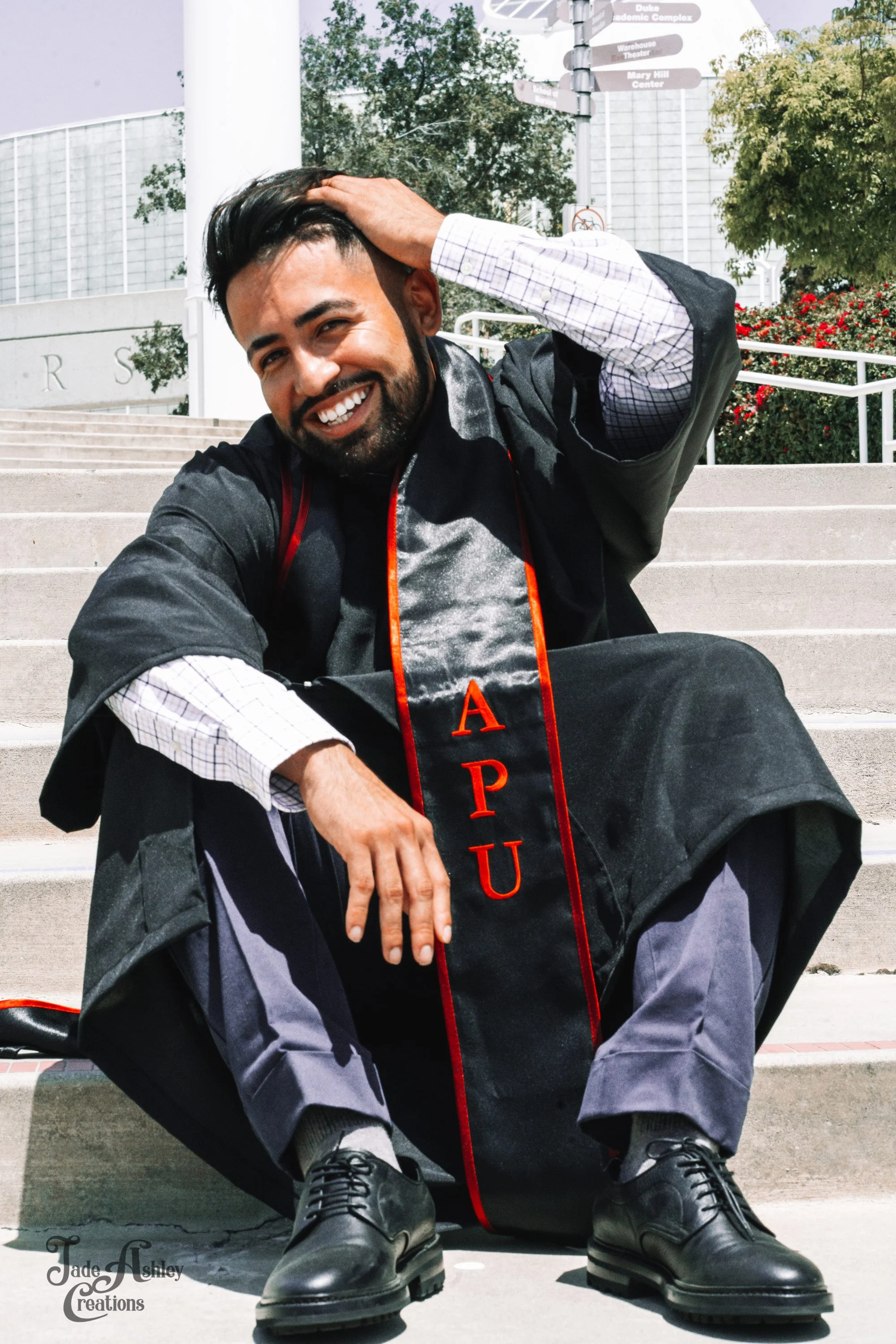 A man in a graduation gown sitting on steps, smiling and running his hand through his hair, with academic buildings and directional signs in the background.