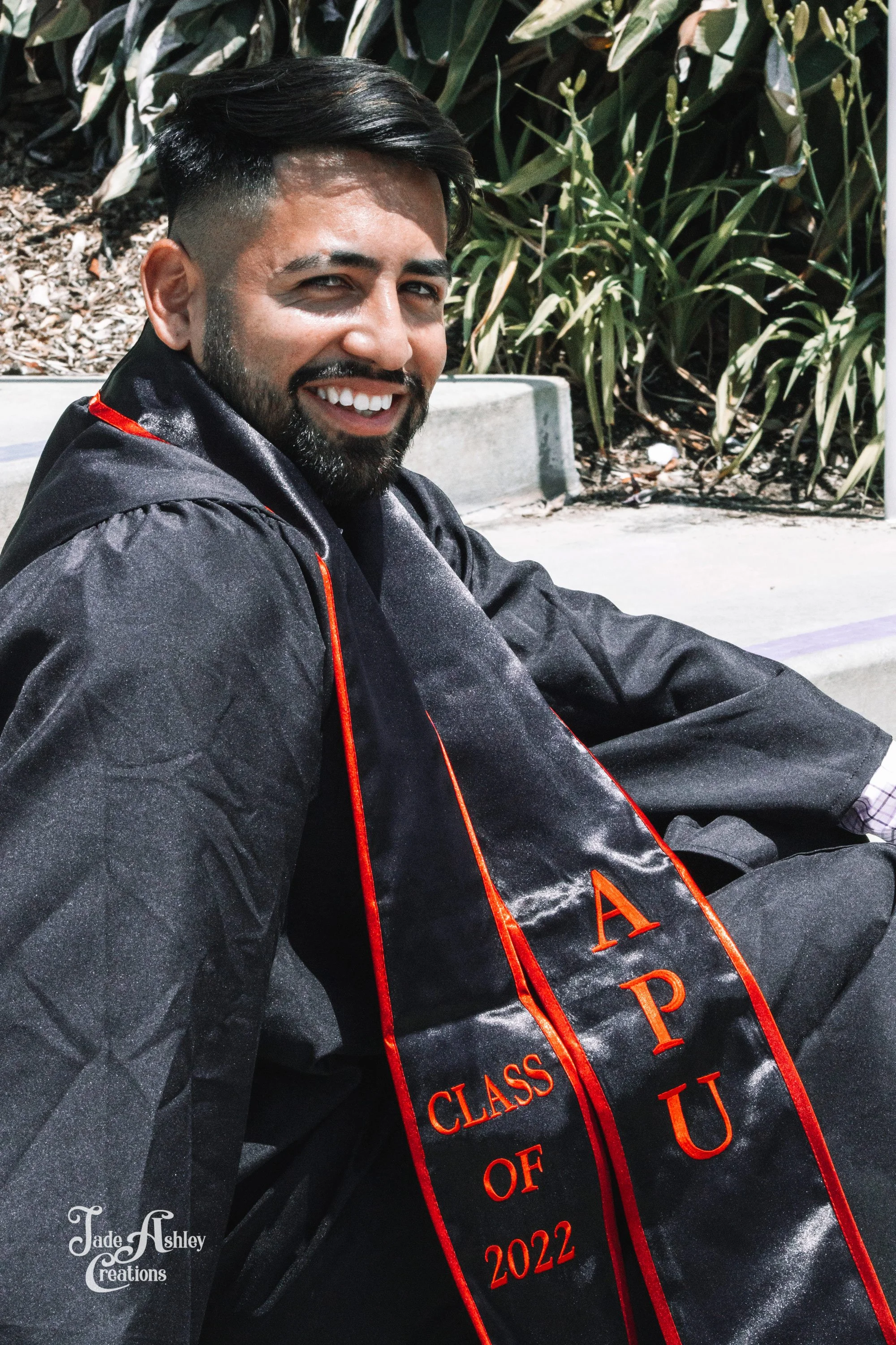 A young man smiling, wearing a graduation gown and stole with 'CLASS OF 2022' and Greek letters, sitting outdoors near greenery.