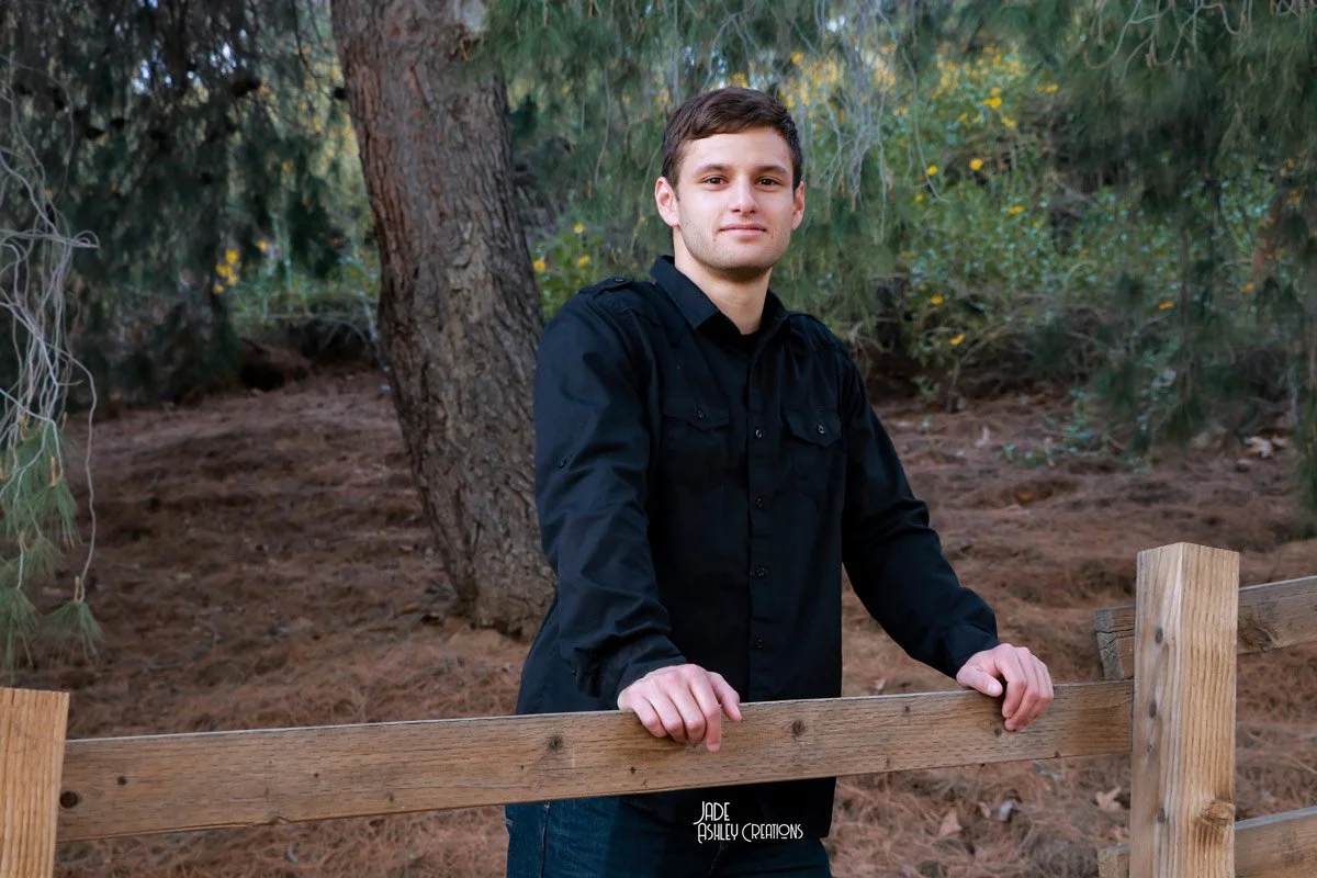 A young man in a black shirt standing outdoors by a wooden fence with trees and foliage in the background.