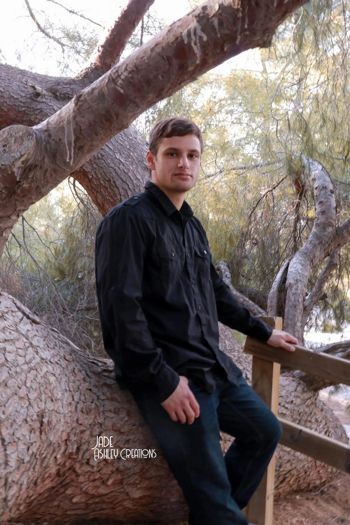 A young man in a black shirt and jeans sitting on a large tree branch outdoors with trees and foliage in the background.