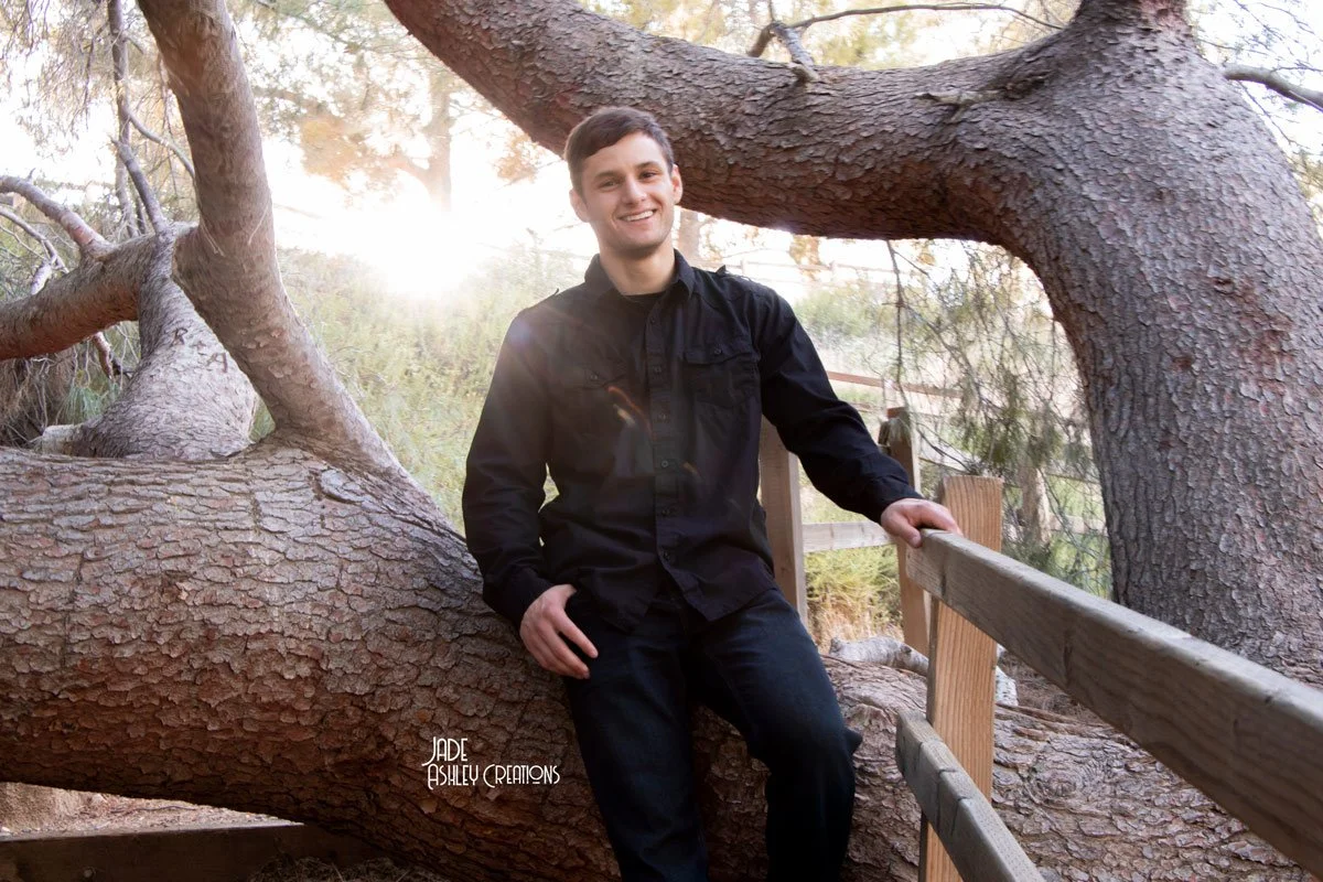 A young man with short brown hair, wearing a black shirt and black pants, sitting on a large, twisted tree branch outdoors during daylight, smiling at the camera with sunlight shining through background trees.
