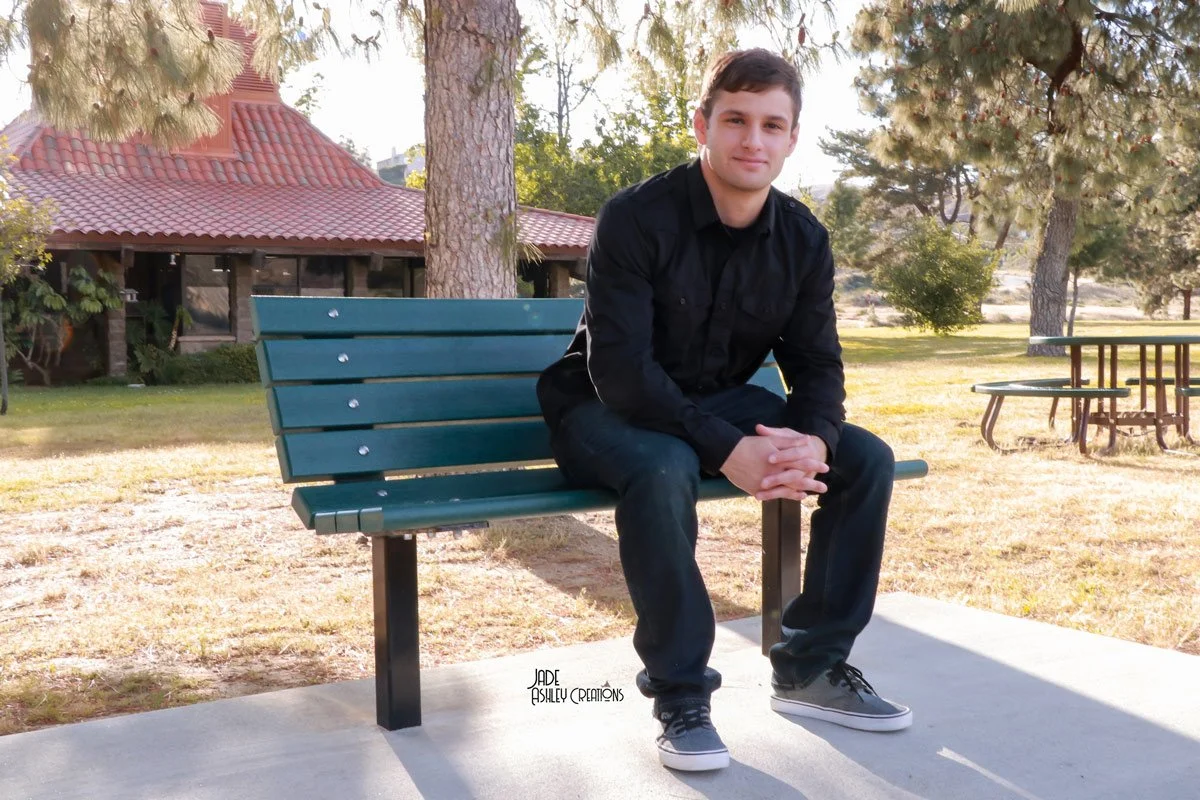 A young man with short brown hair, wearing a black button-up shirt, dark jeans, and black sneakers, sitting on a green park bench outdoors with trees and a building in the background.