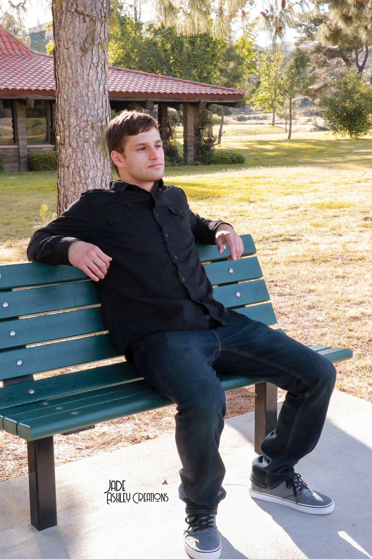 A young man with brown hair, wearing a black shirt, jeans, and sneakers, sits relaxed on a green park bench under a large tree in a sunny park with grass, trees, and a building in the background.