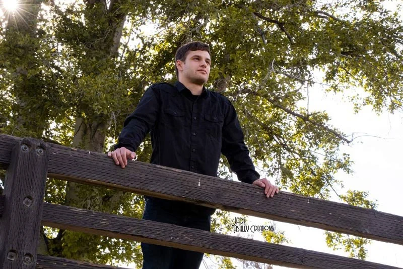 Young man with dark hair in a black shirt leaning on a wooden fence outdoors with trees and green foliage in the background.