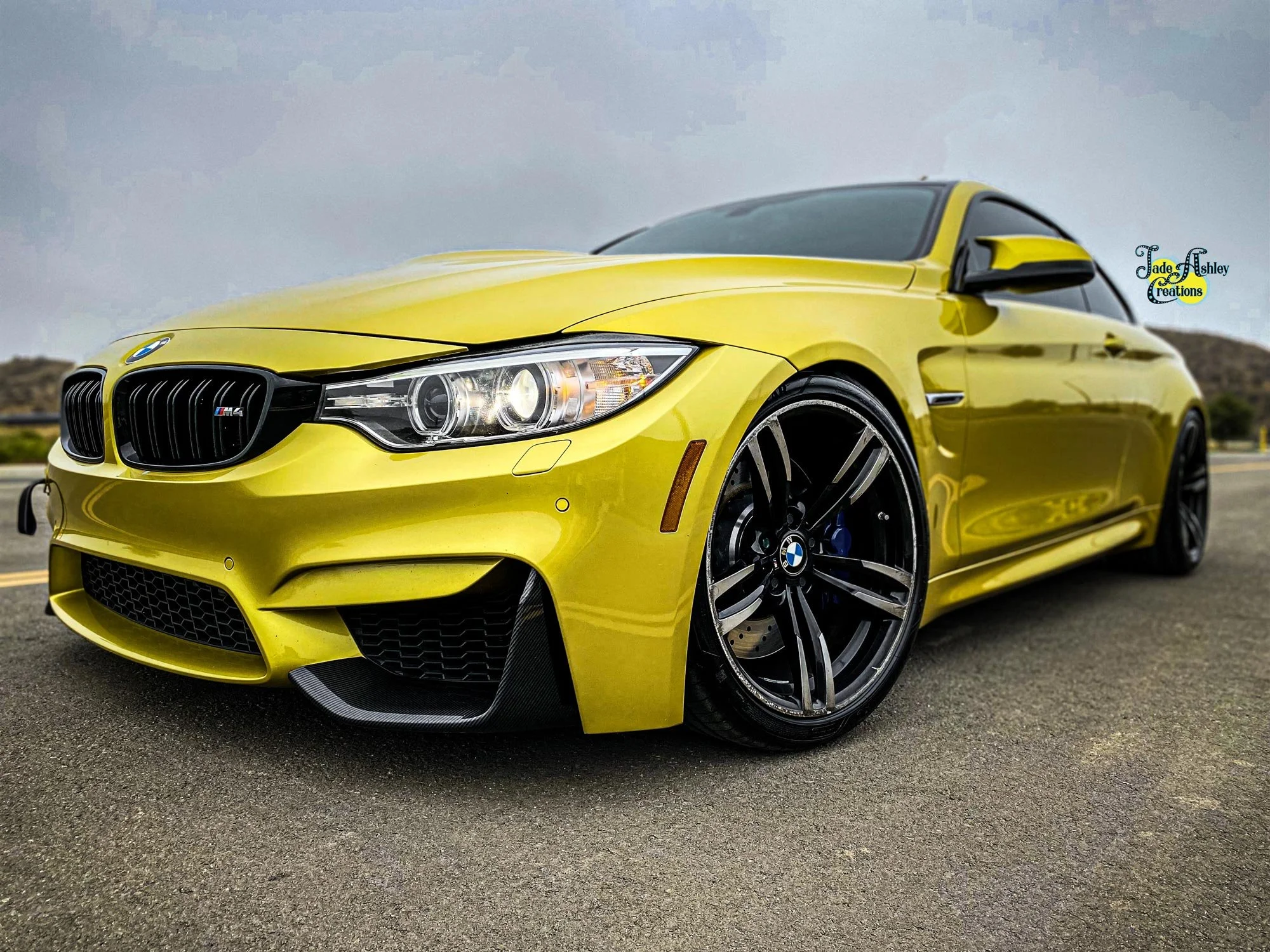 A bright yellow BMW M4 sports car parked on a paved surface with a cloudy sky and distant hills in the background.