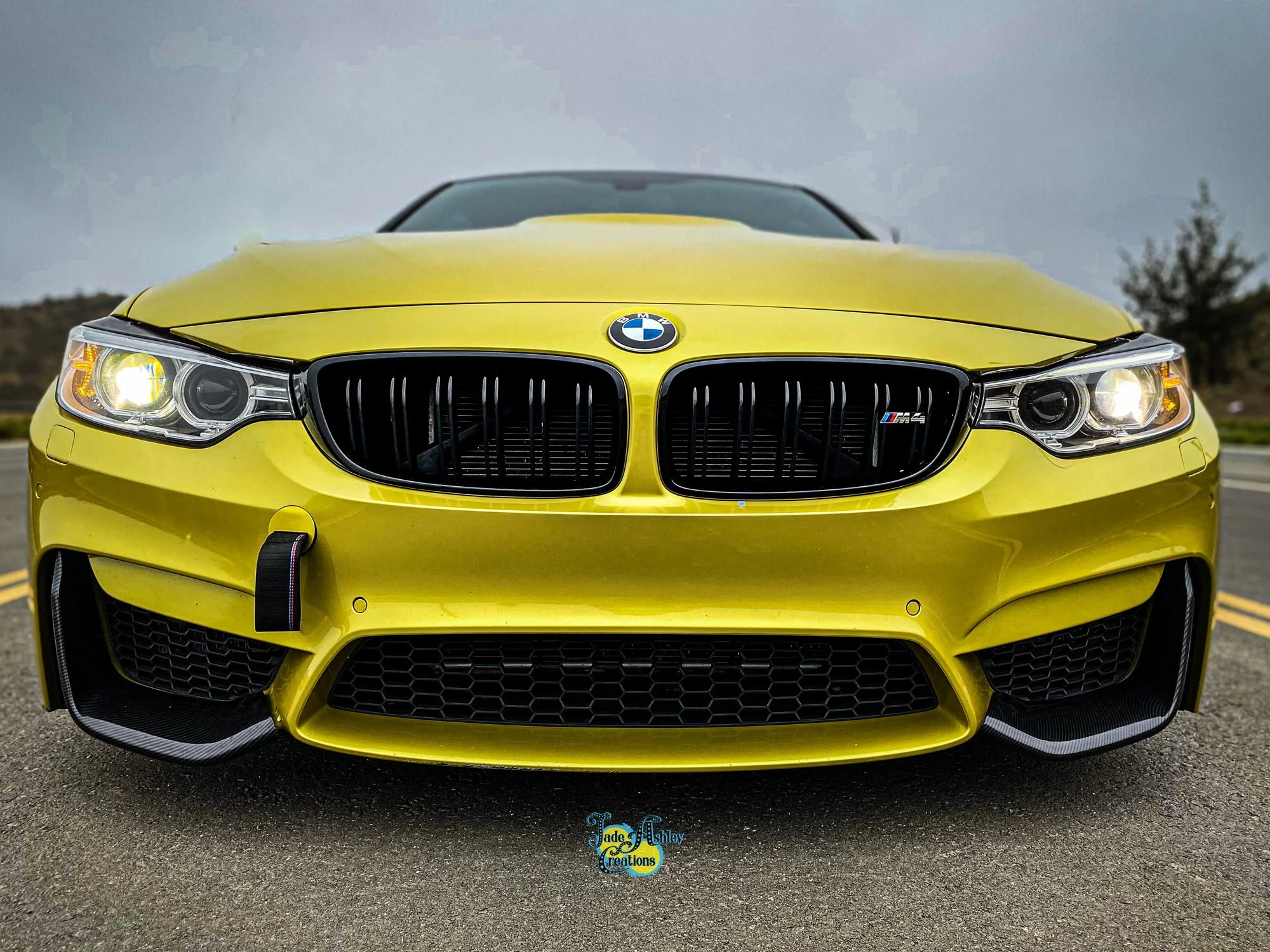 Front view of a yellow BMW M4 with black grille and sporty design, parked on an asphalt road under cloudy sky.