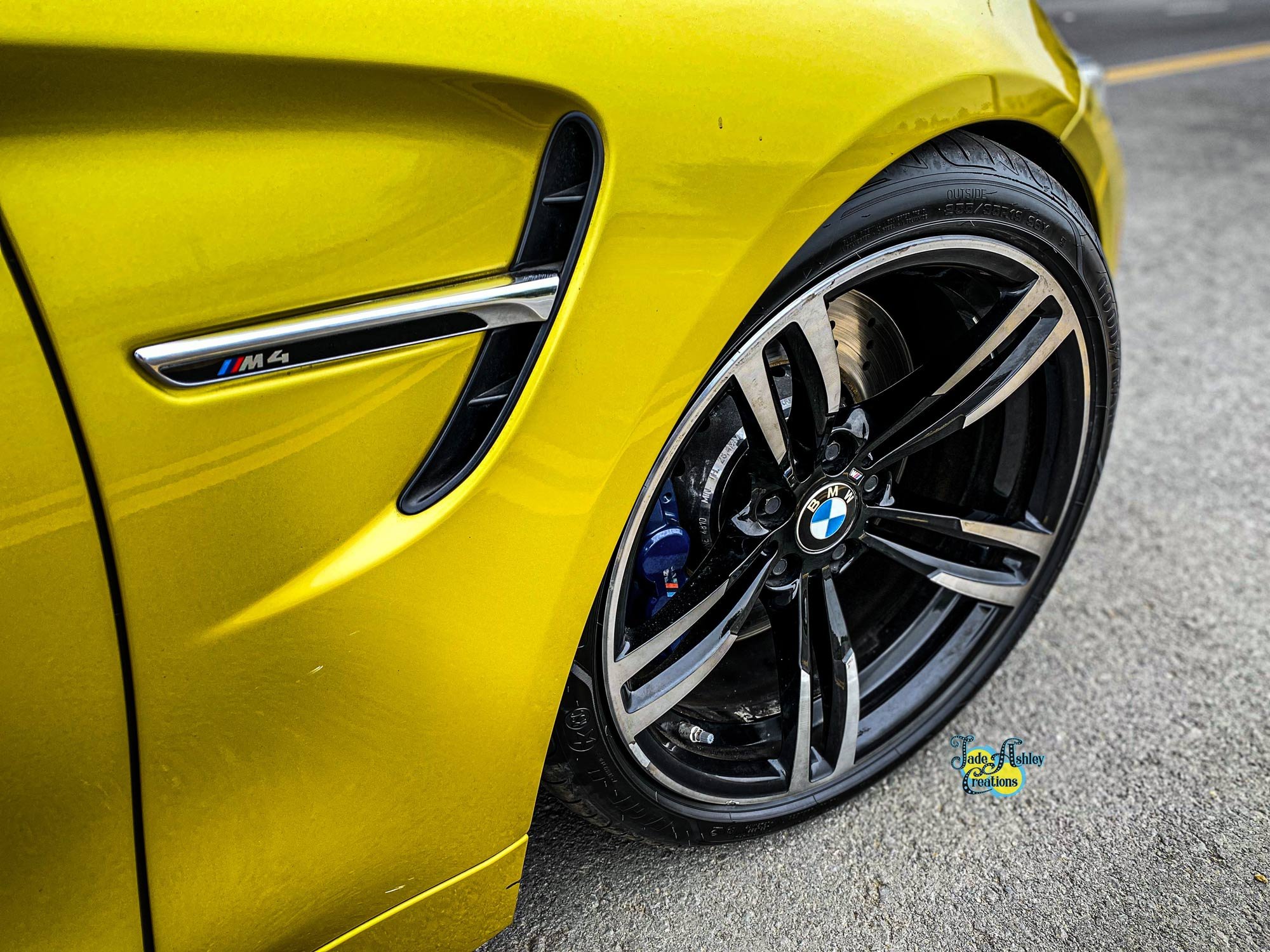 Close-up of the front side of a bright yellow BMW M4 car, showing the wheel, rim, and side vent with M4 badge.