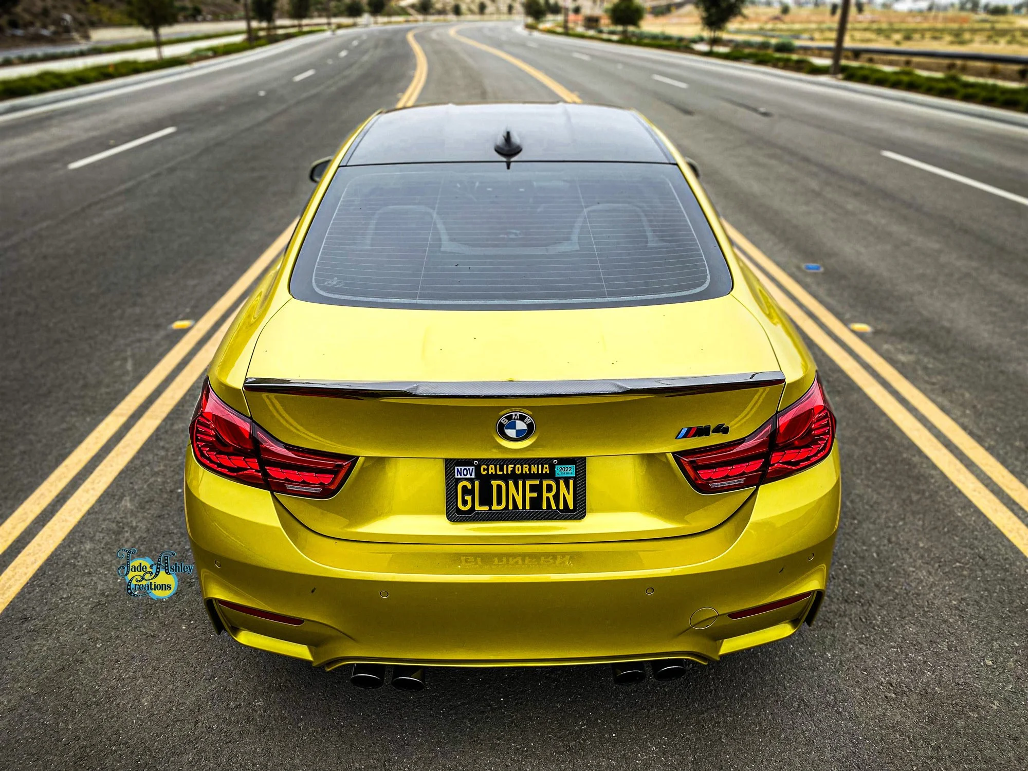 Yellow BMW M4 sports car with California license plate GLDNFN, parked in the middle of a multi-lane road with yellow double lines, under a clear sky.