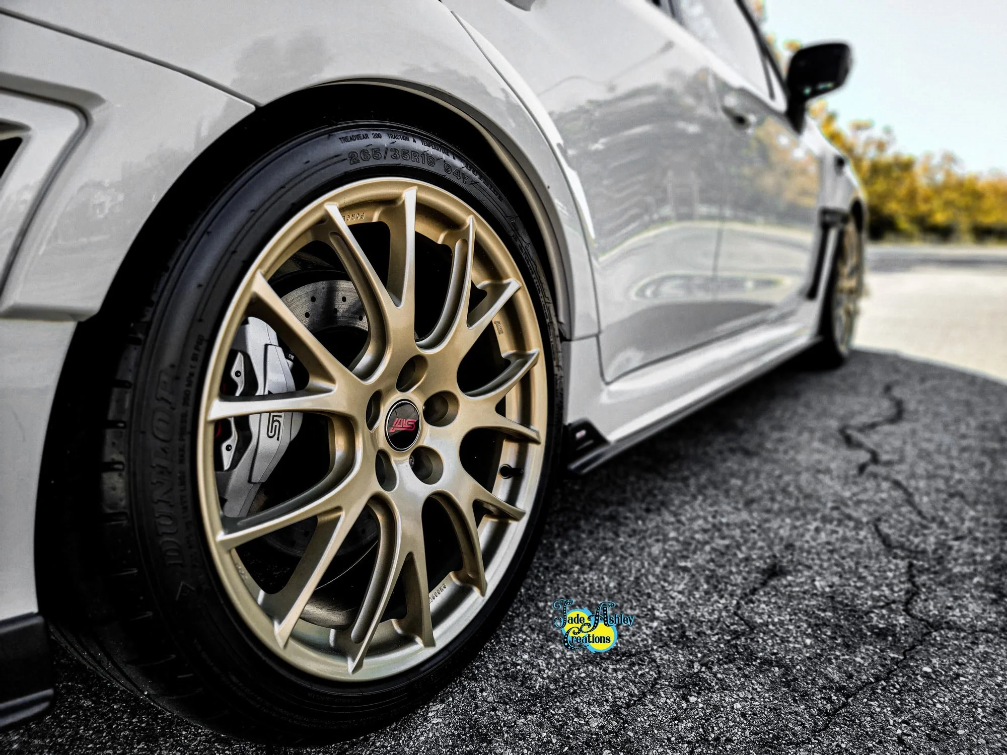 Close-up of a silver sports car's front wheel with gold alloy rims and a high-performance tire, parked on a textured asphalt surface with a blurred background of trees.
