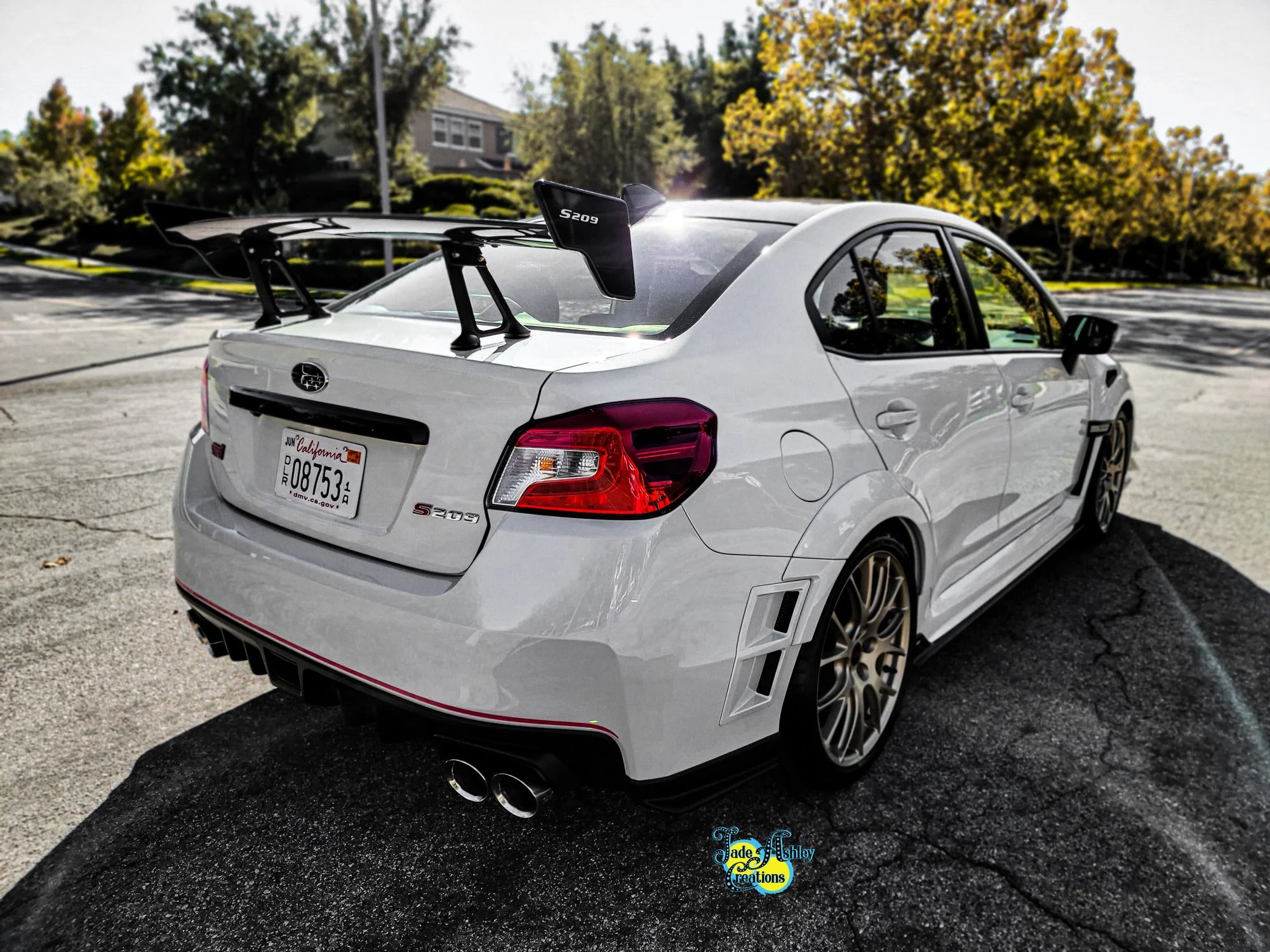 A white race car with a prominent rear spoiler, aftermarket wheels, and dual exhaust pipes parked on a street in a residential neighborhood with trees and houses in the background.