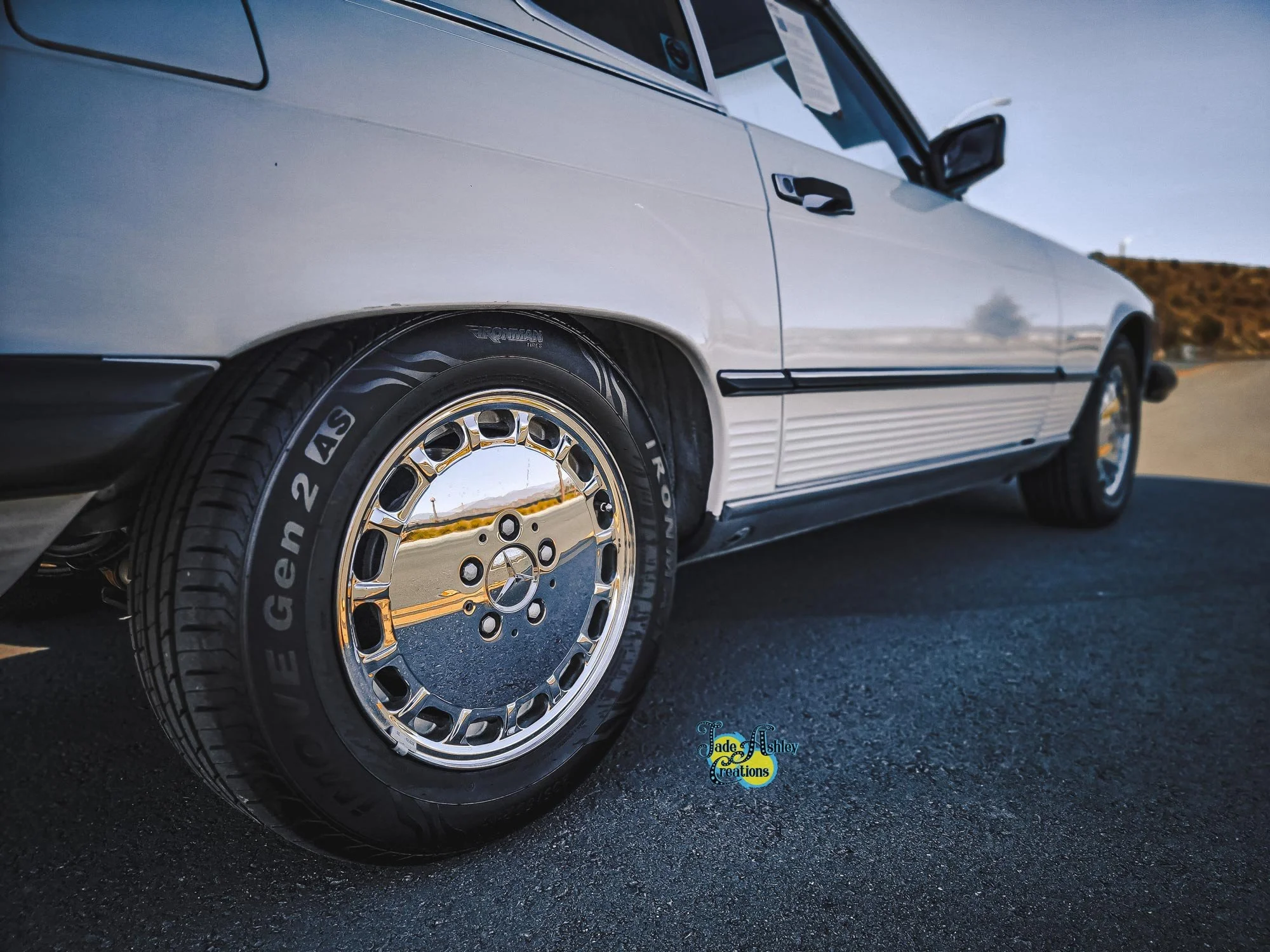 Close-up of a vintage silver Mercedes-Benz parked on an asphalt road, showing front wheel with reflective rim and branded tire.