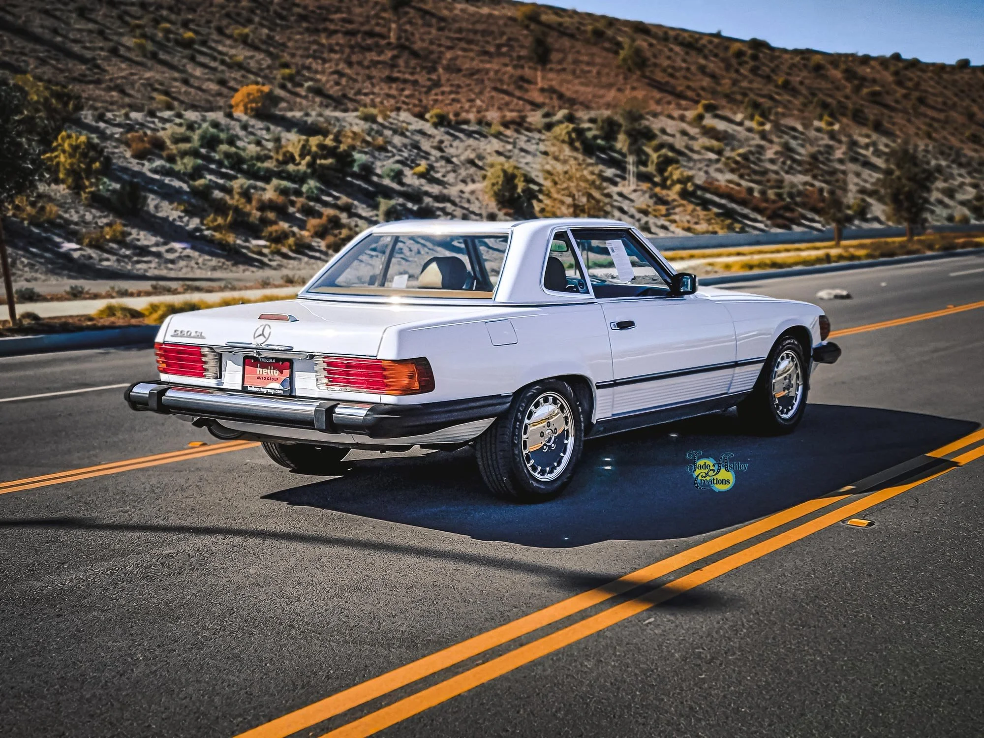 A white vintage Mercedes-Benz 560 SL parked on a deserted highway with desert hills in the background.
