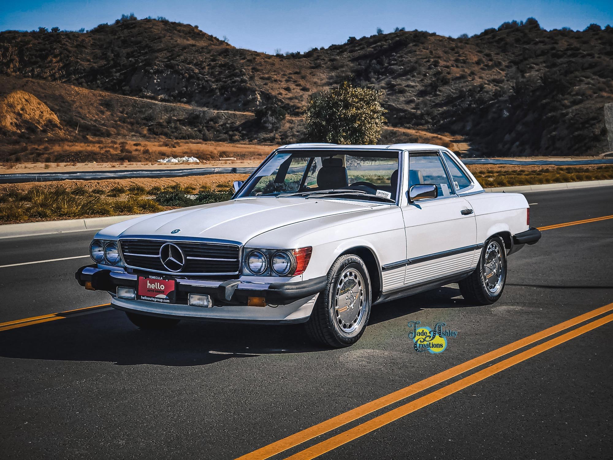 A vintage white Mercedes-Benz coupe parked on a desert highway with mountains in the background.