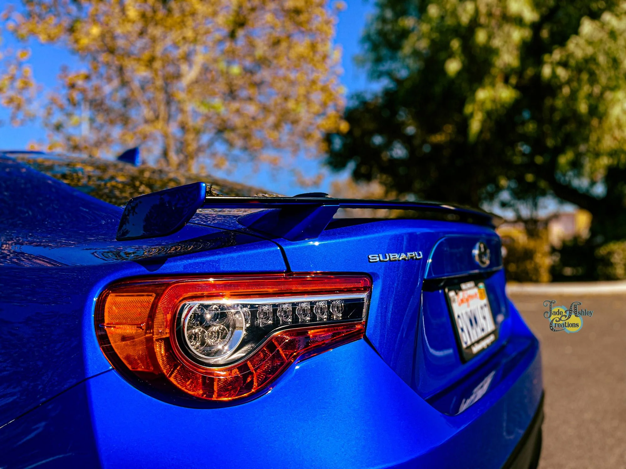 Close-up of the rear of a blue Subaru car, showing the tail light, spoiler, and license plate, with trees and a clear sky in the background.