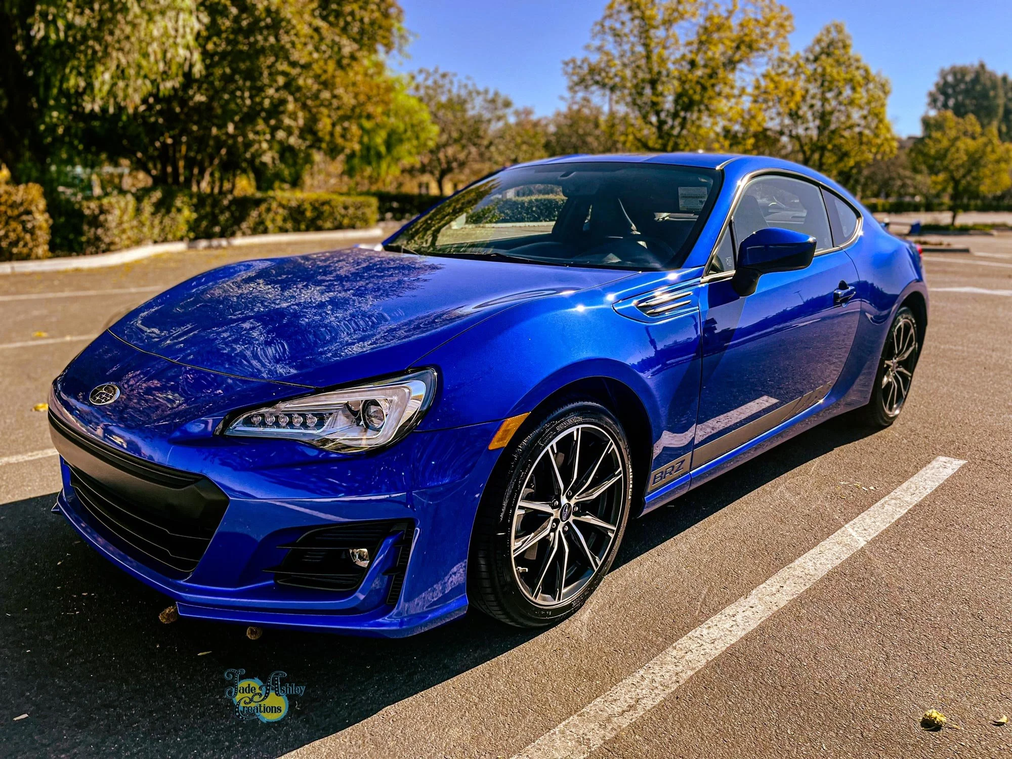 A blue Subaru BRZ sports car parked in an outdoor parking lot, with trees and blue sky in the background.