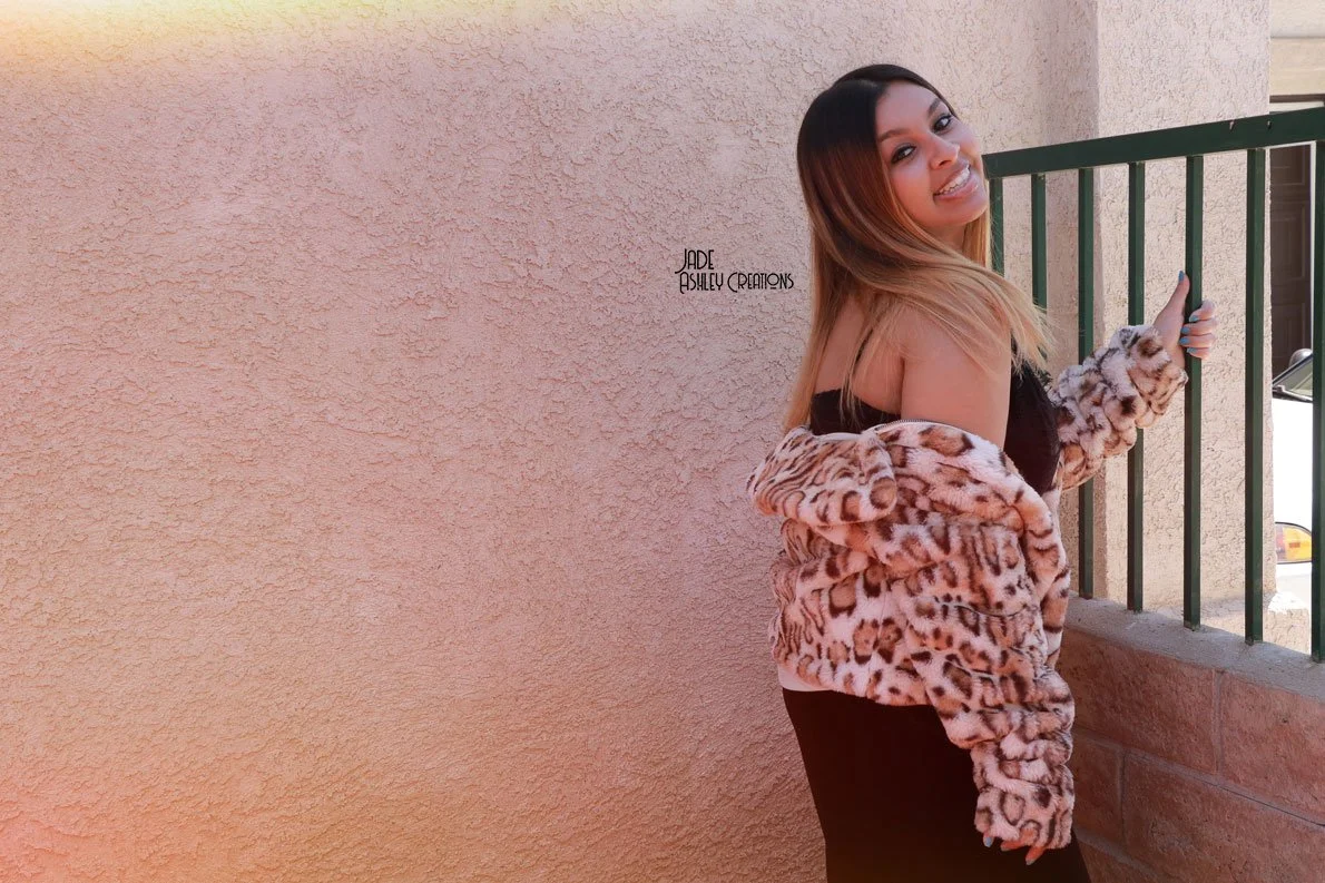 A woman in a leopard print faux fur jacket, black top, and black pants standing next to a green metal railing on a balcony with beige stucco walls in the background, smiling and slightly tilting her head.