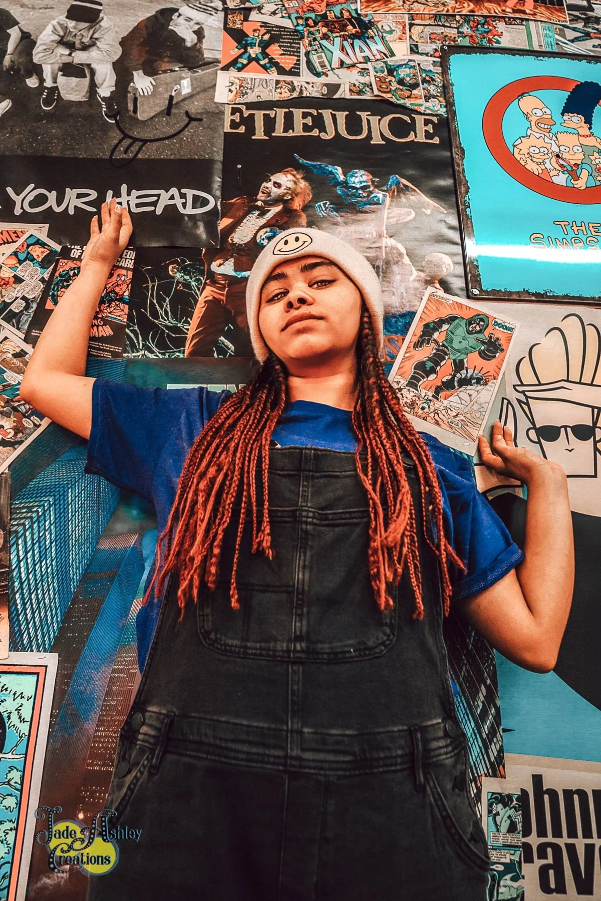 Teen girl with orange dreadlocks wearing a gray beanie with a smiley face, denim shirt, and overalls, standing in front of a wall decorated with various comic book posters and artwork.