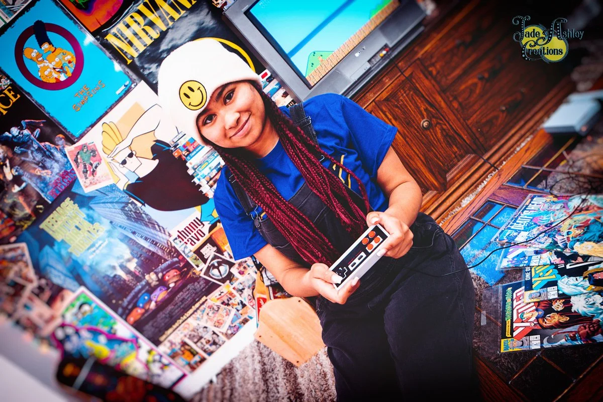 A young girl with braided red hair wearing a white beanie with a smiley face patch, smiling while holding a gaming controller in a room decorated with comic book posters and artwork.