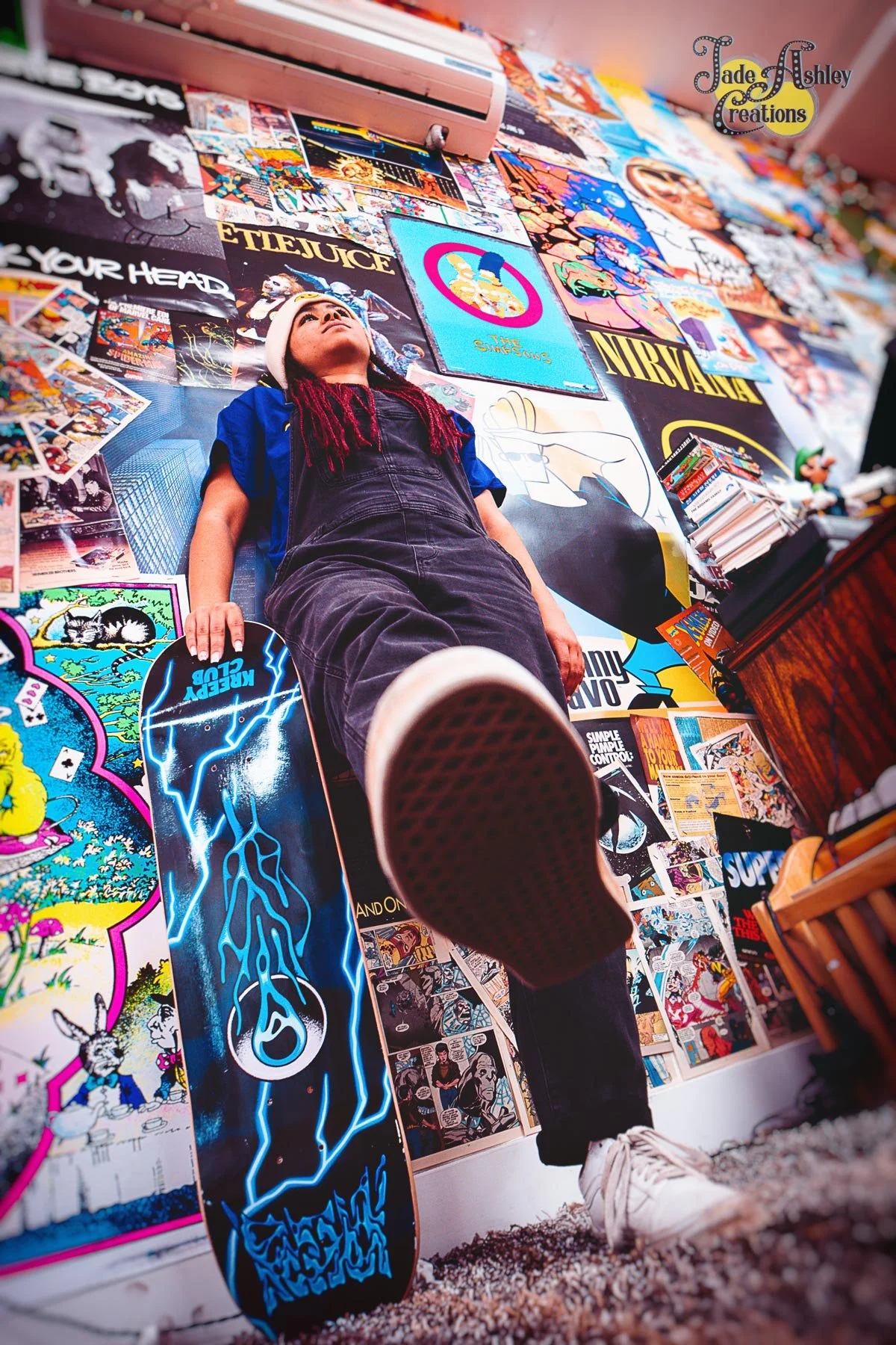 A person with dreadlocks wearing a blue shirt and black overalls is lying on a bed or floor, with their head tilted back and feet toward the camera. They are holding a skateboard with lightning graphics. The background wall is covered with posters an